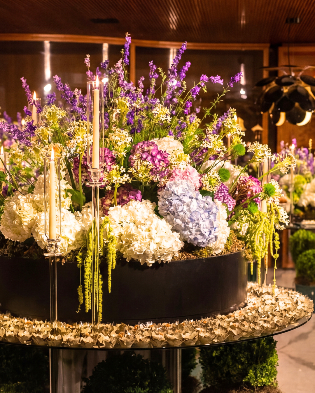 Arranjo de flores em tons de roxo, branco e verde com velas acesas em um ambiente interno decorado.