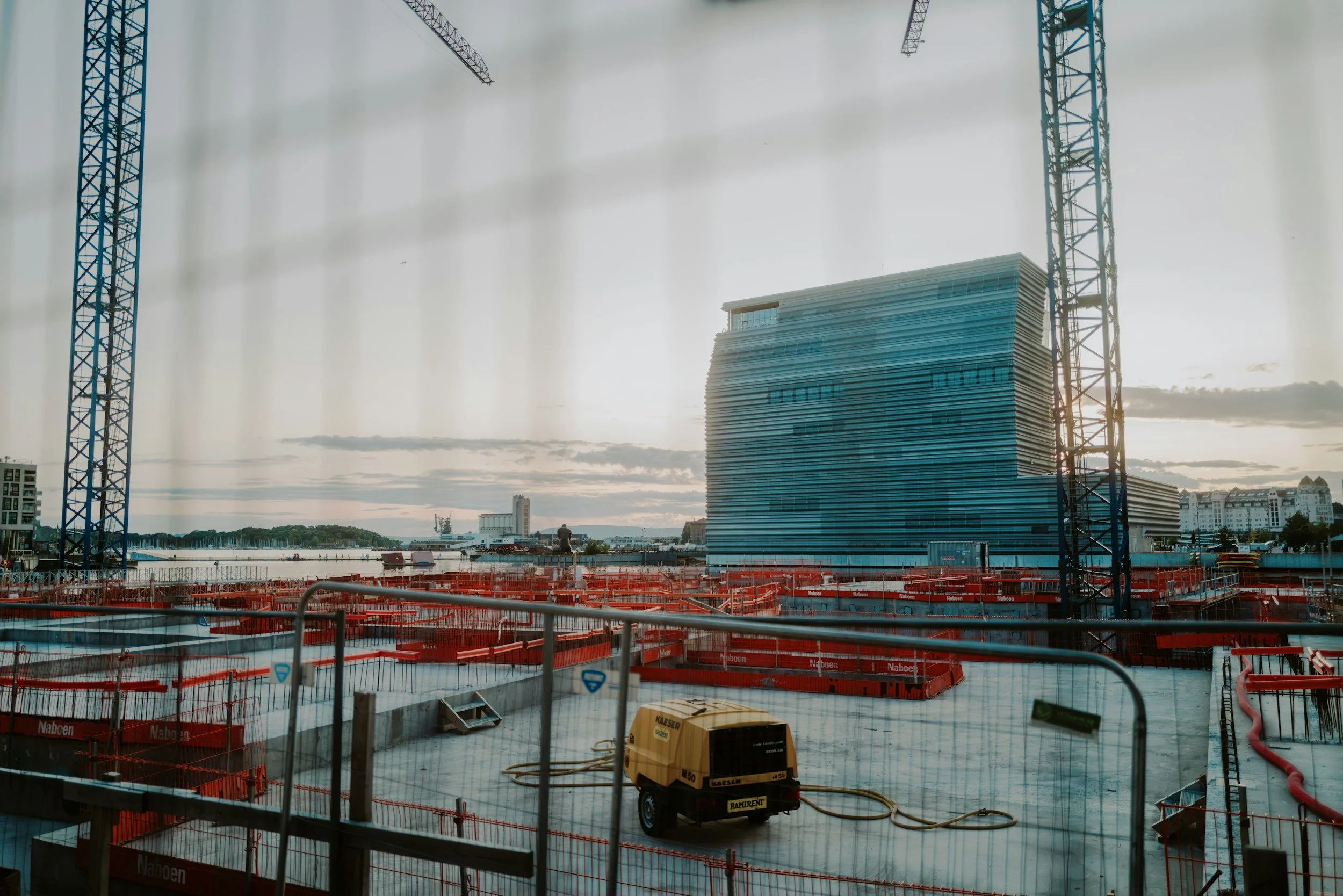 Byggeplass med skyskraper under konstruksjon, store kraner og utstyr, og en by i bakgrunnen, sett gjennom et gitter.