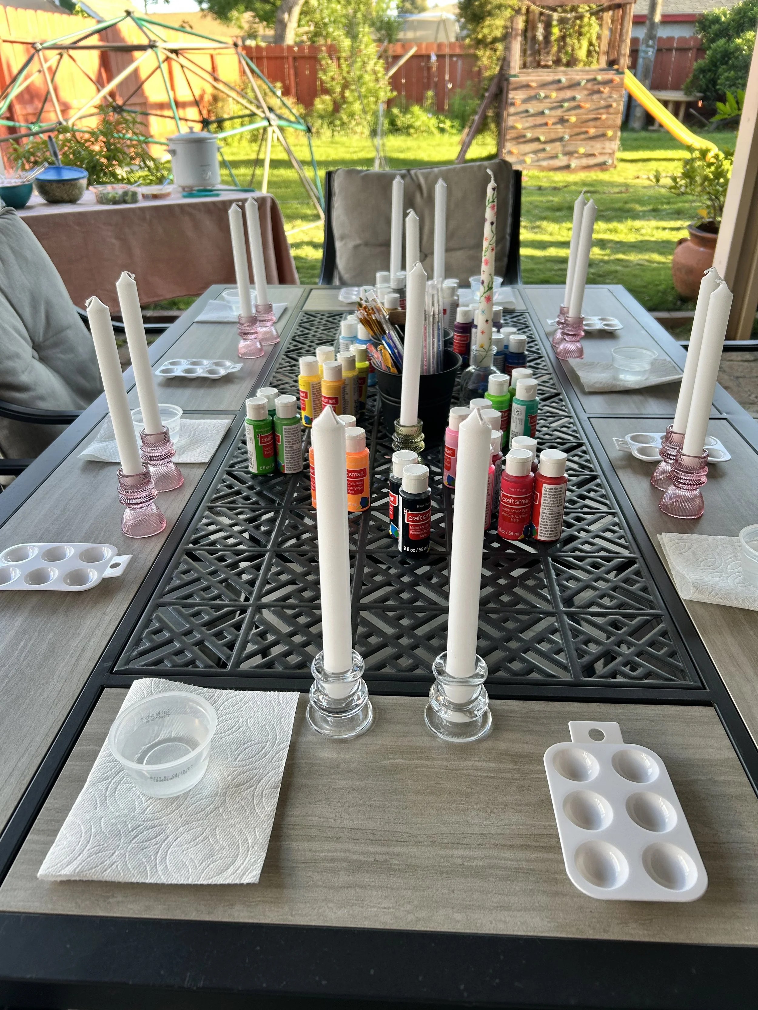 table set with taper candles in candleholders, paint bottles, paint pallettes, paper towels, cups, and paint brushes in small bucket