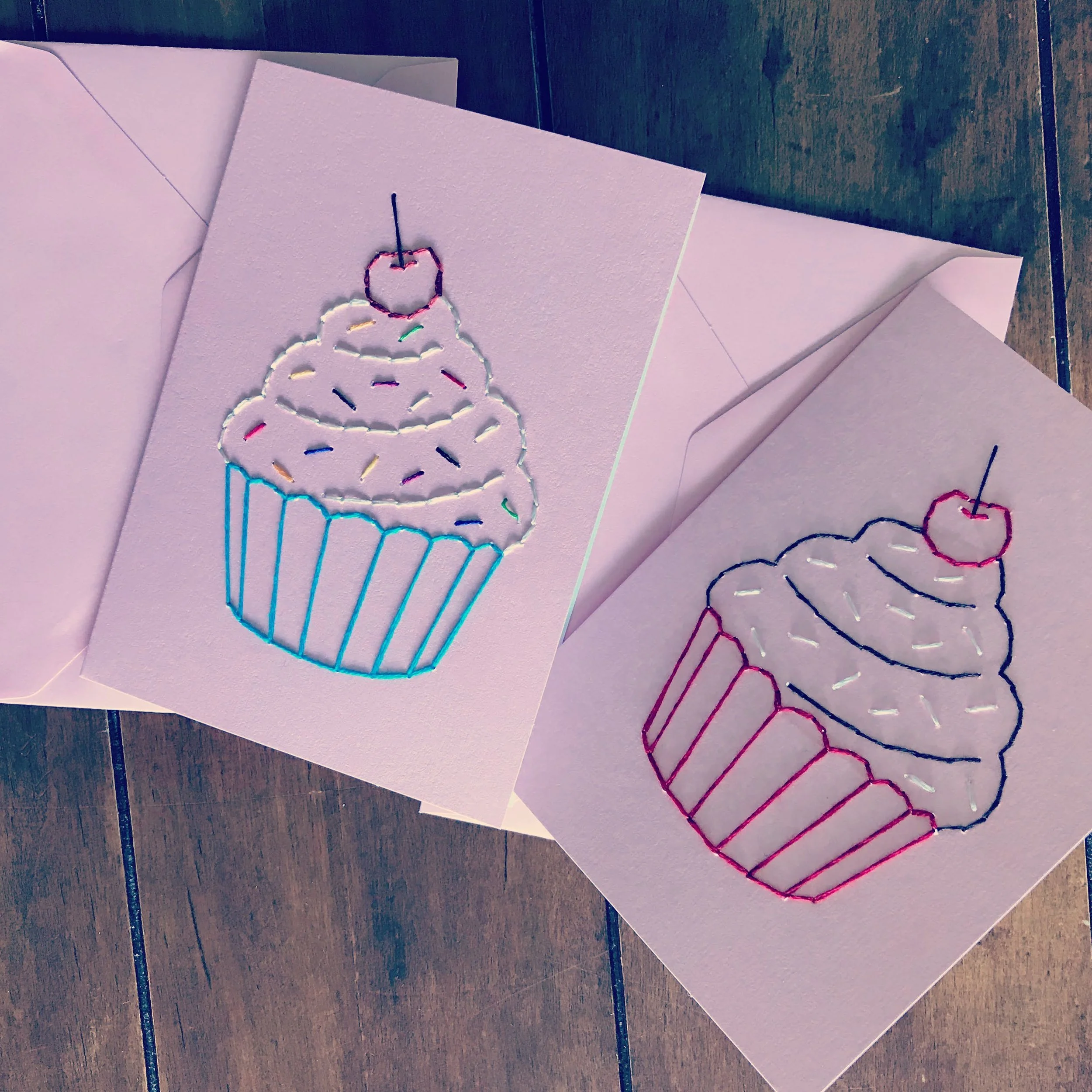 two pink cards with colorful embroidered cupcake silhouettes nested inside pink envelopes atop wood boards