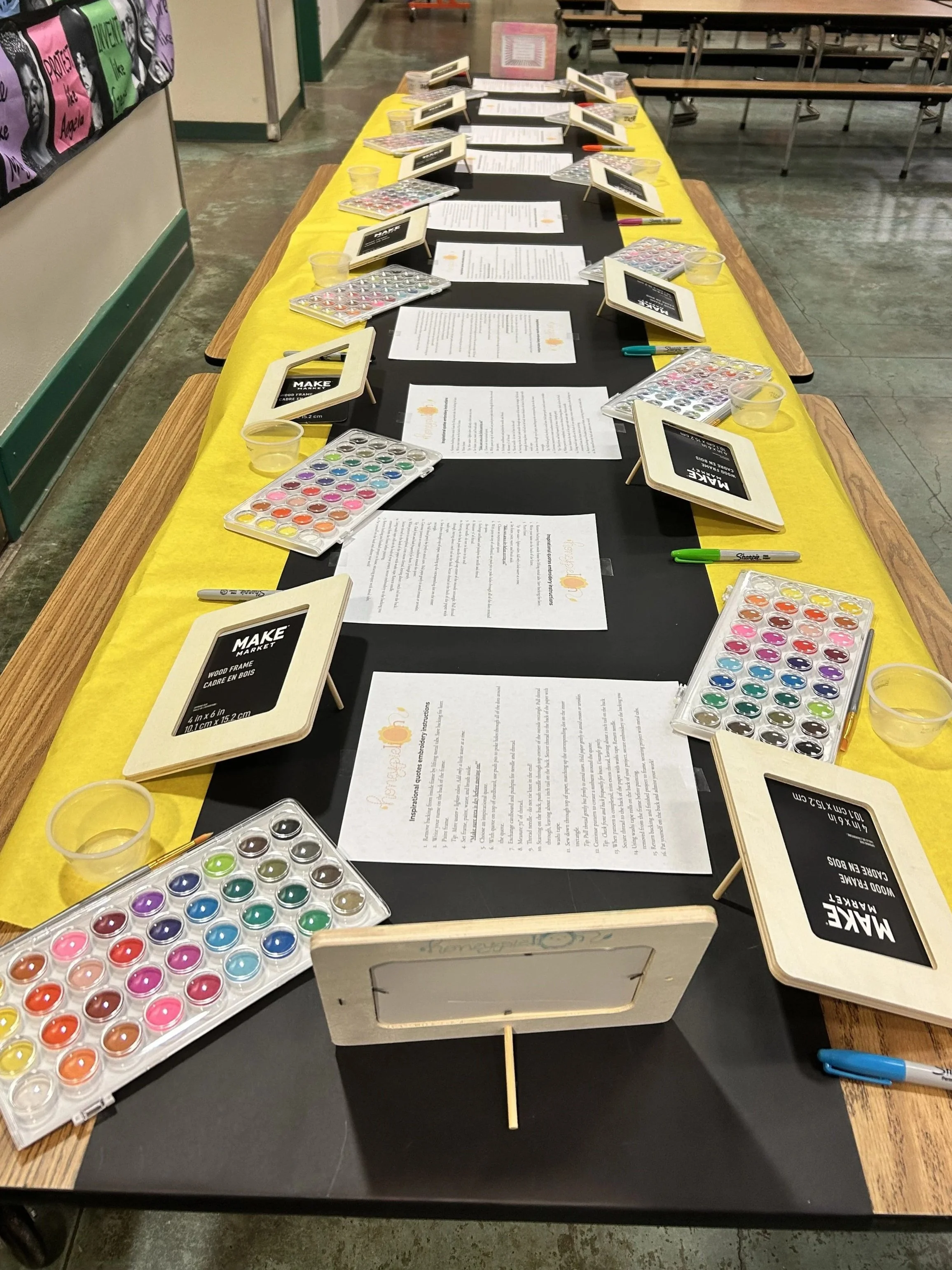 table covered in yellow and black paper runners,  with wood frames, watercolor palette, markers, cups, and paper instructions