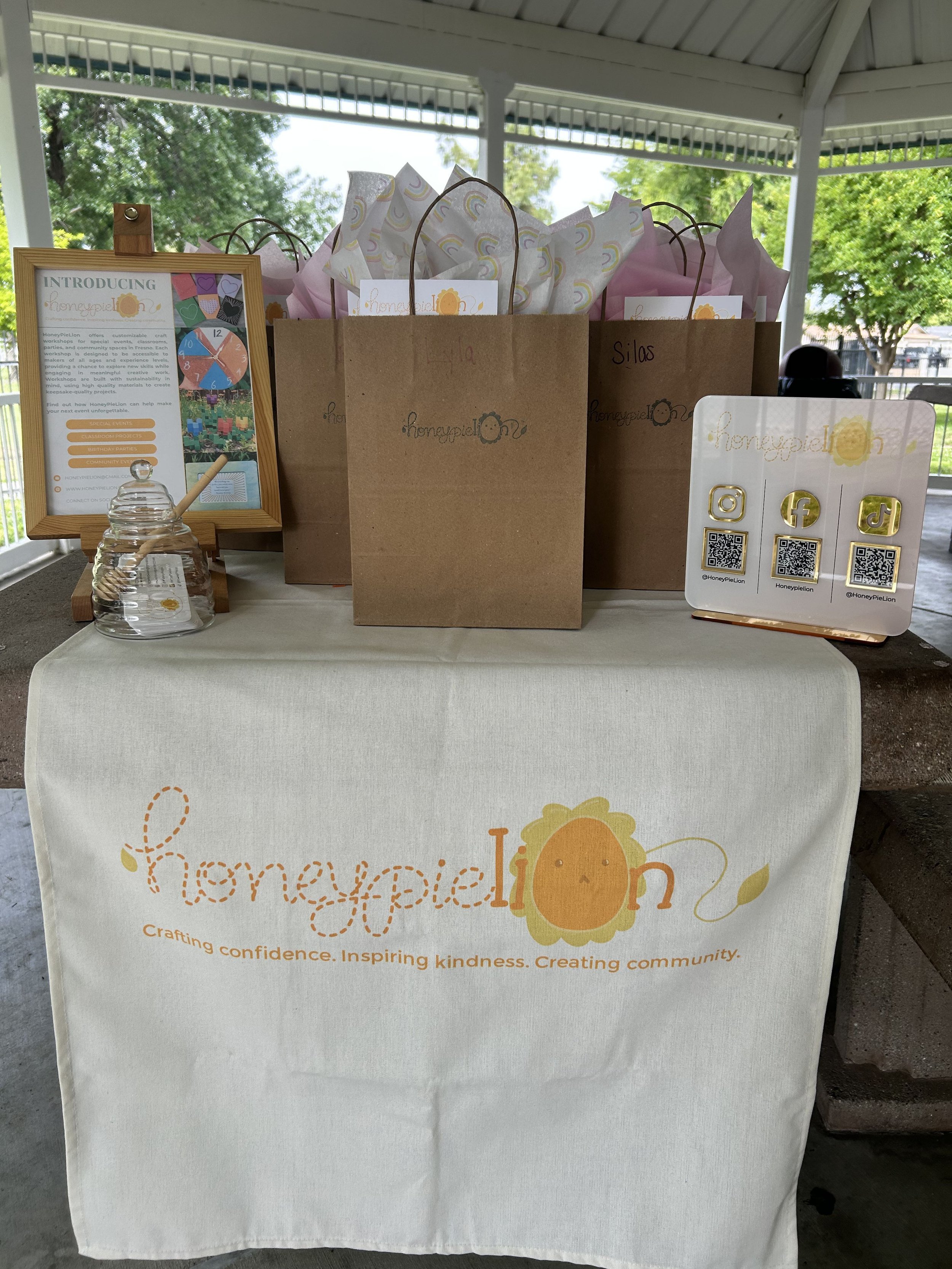brown paper bags with tissue paper on top of HoneyPieLion branded table runner, featuring honeypot business card holder, qr codes for social media links, and "about HoneyPieLion" flyer