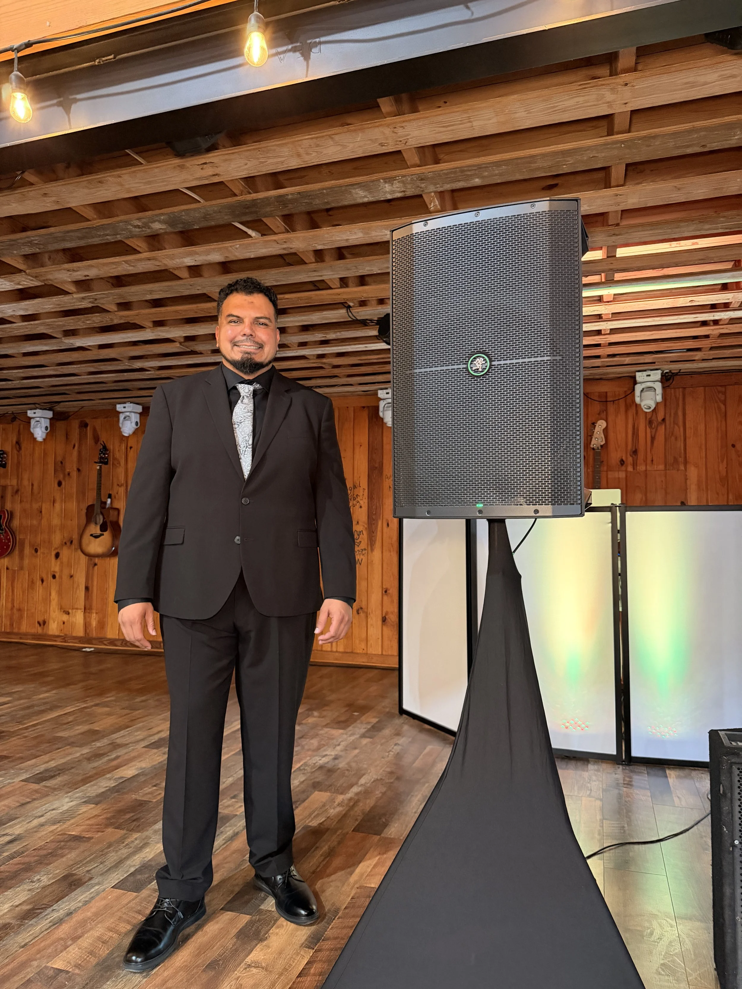 A man in a black suit and silver tie standing next to a large black speaker on a stand at an event with wooden walls and ceiling, and guitar decorations in the background.