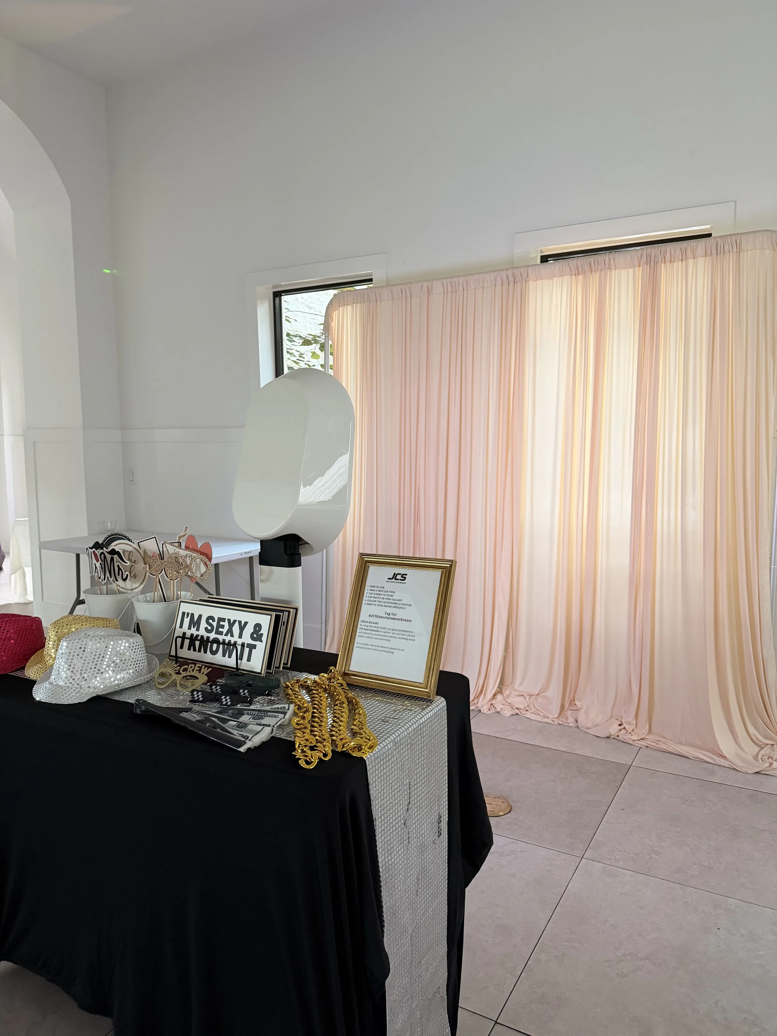 Party photo booth with accessories like hats, signs, and gold chains, set up against pink curtains.