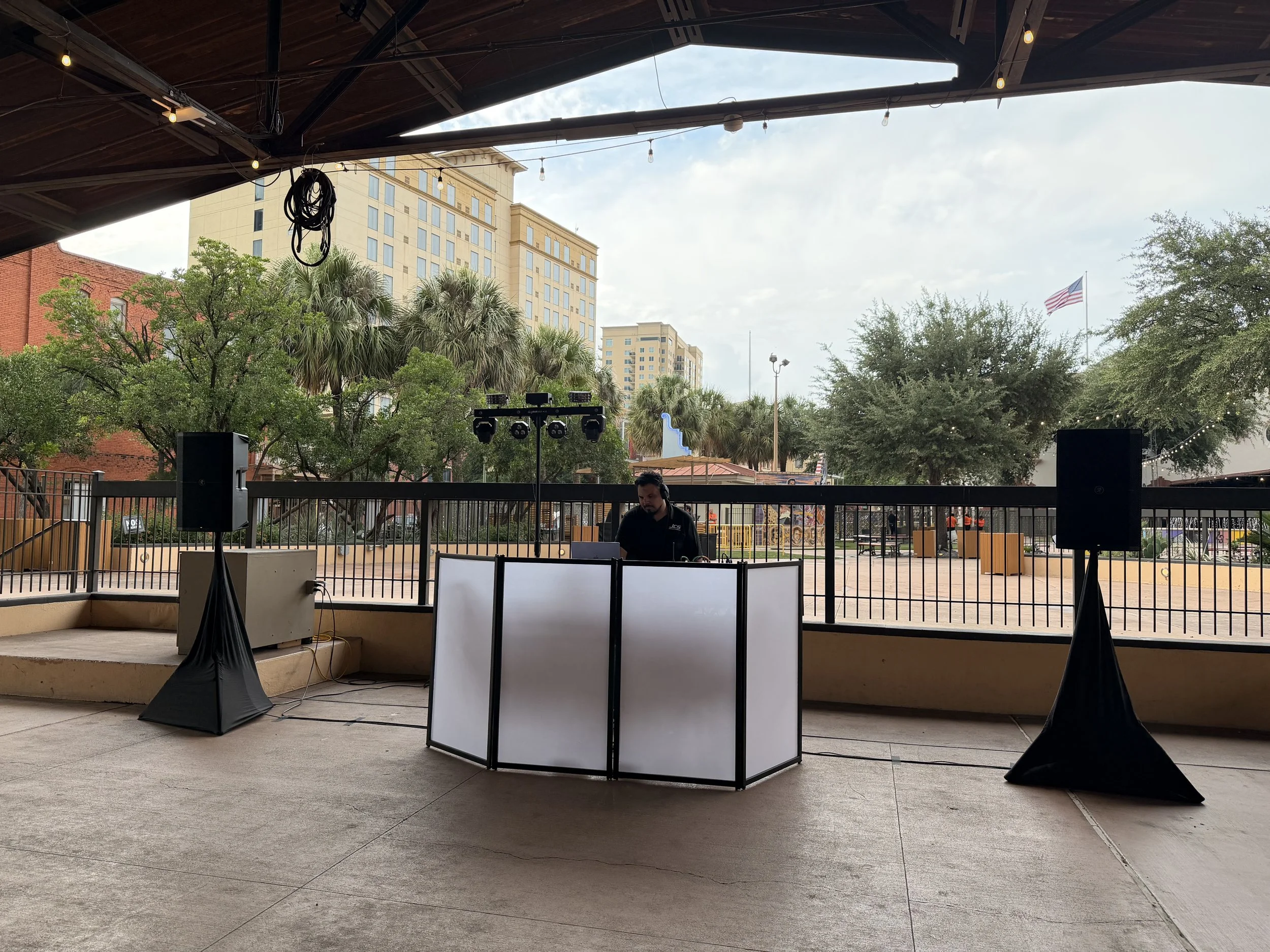 Empty outdoor stage with sound equipment, a person working, trees, tall buildings, and an American flag in the background.