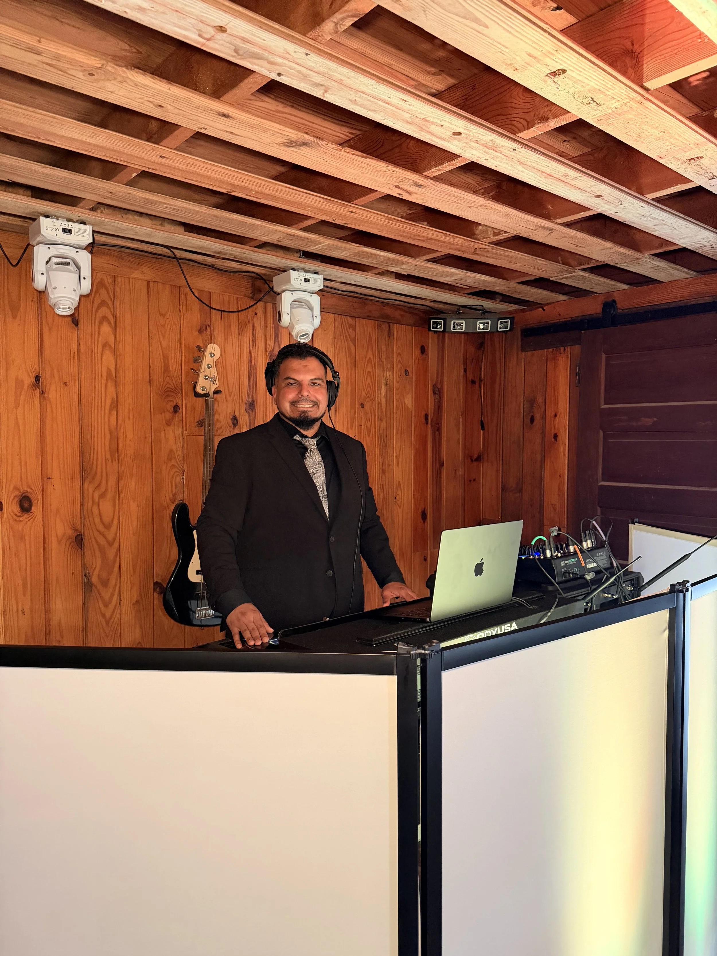 A man in a suit wearing headphones, standing behind a DJ or music setup, smiling at the camera in a wooden-paneled room.