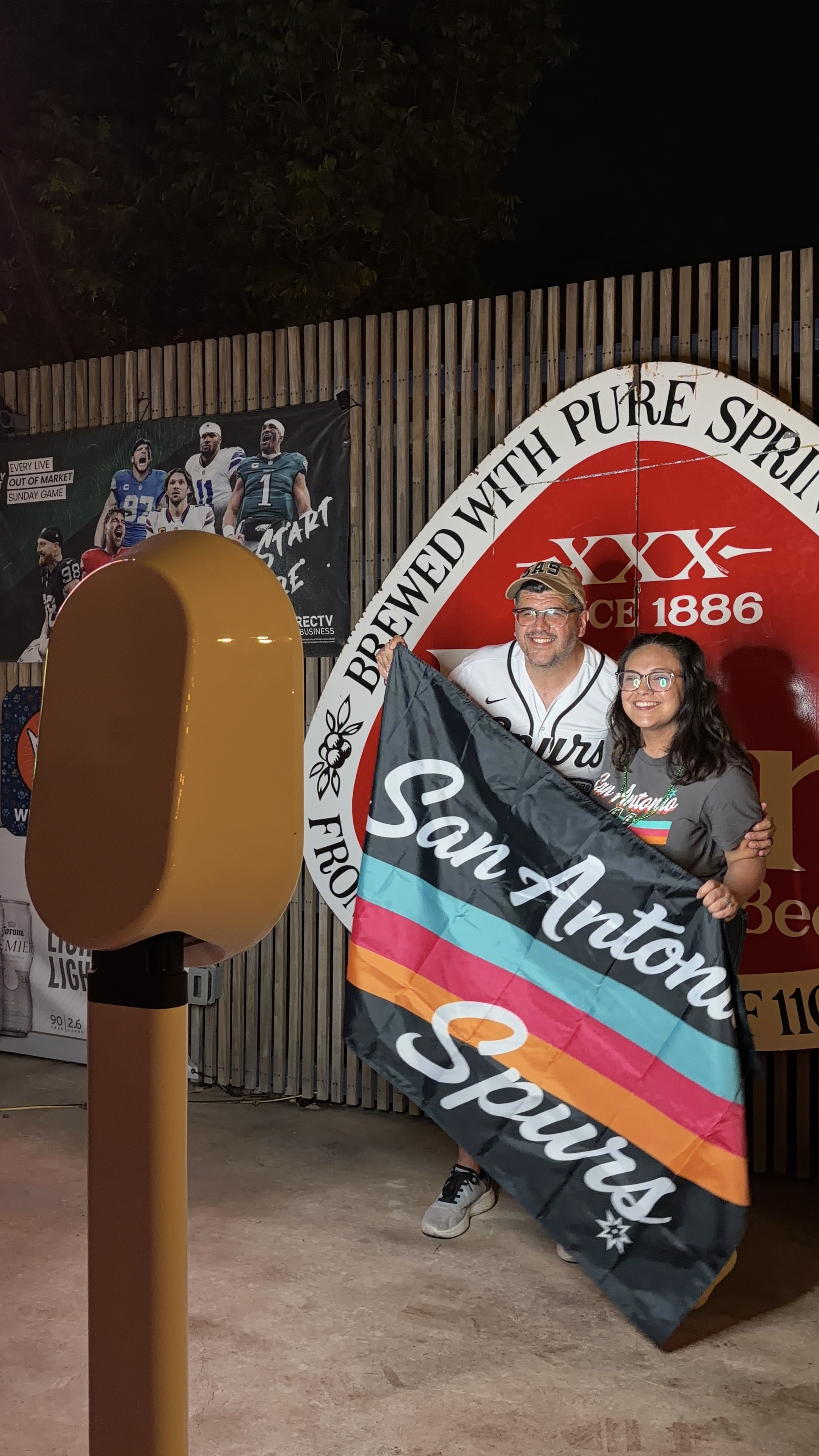 Two people posing with a pride flag in front of a large image of football players and a circular sign with the text "Brewed with pure spirit." One person holds a pride flag, and they are smiling. There is also a large yellow beer tap in the foregroun