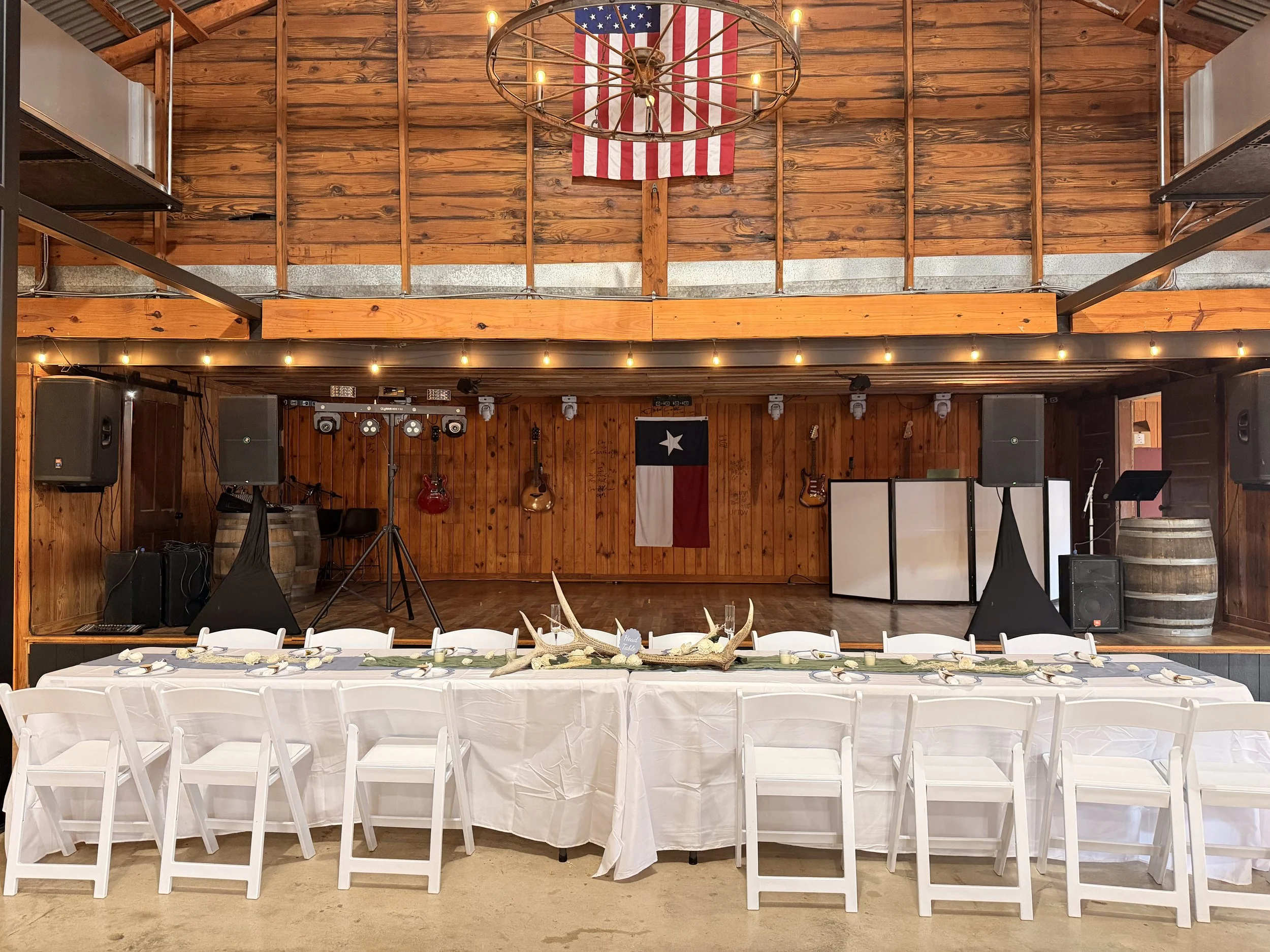 A rustic indoor venue with wooden walls and ceiling, decorated for an event with a long table covered in a white tablecloth, set with plates and silverware. The table is surrounded by white chairs and decorated with antlers and candles. In the backgr