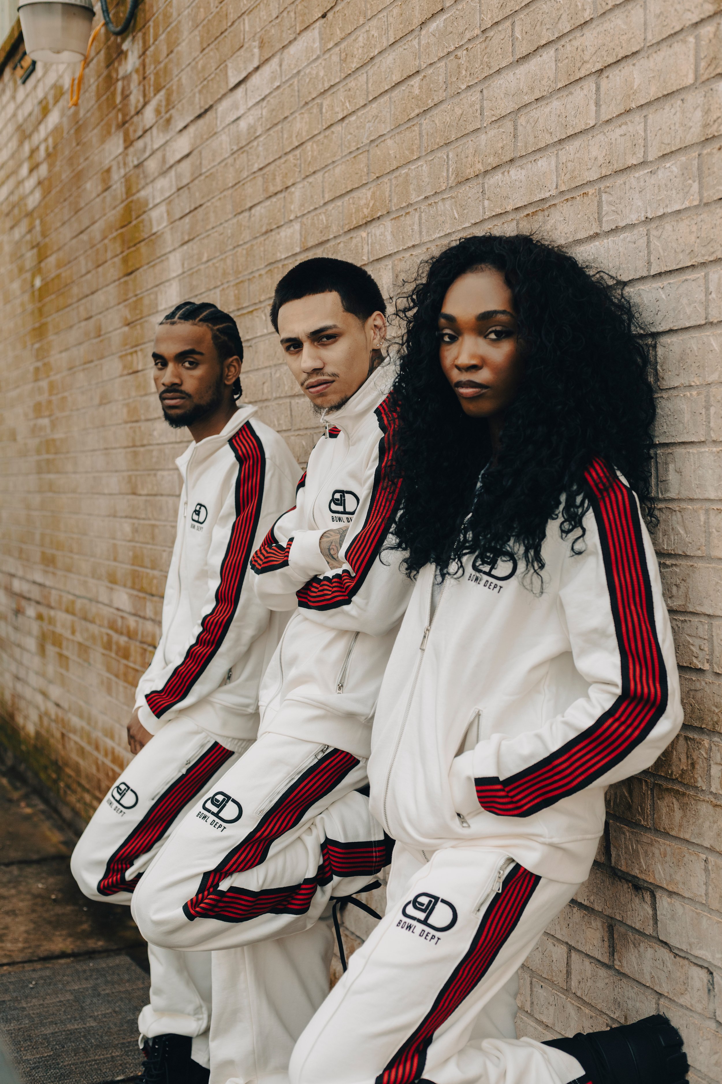 Three young adults wearing matching white tracksuits with red and black stripes, standing against a brick wall. The tracksuits have a logo that reads 'Bowl Dept'. The individuals have serious expressions and are looking at the camera.