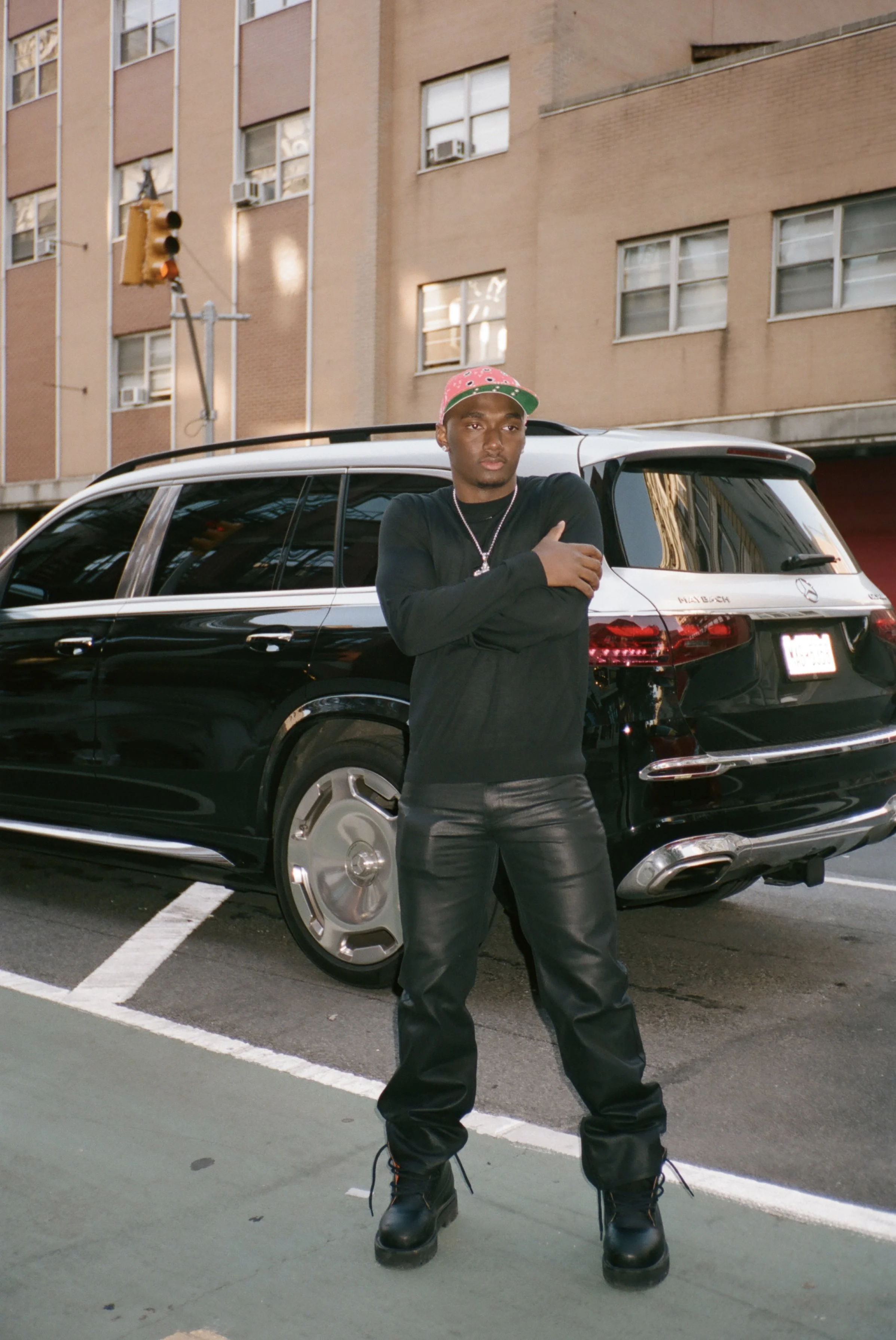 Young man standing in front of a black SUV in an urban parking lot, wearing a pink and green baseball cap, black long sleeve shirt, black leather pants, and black boots.