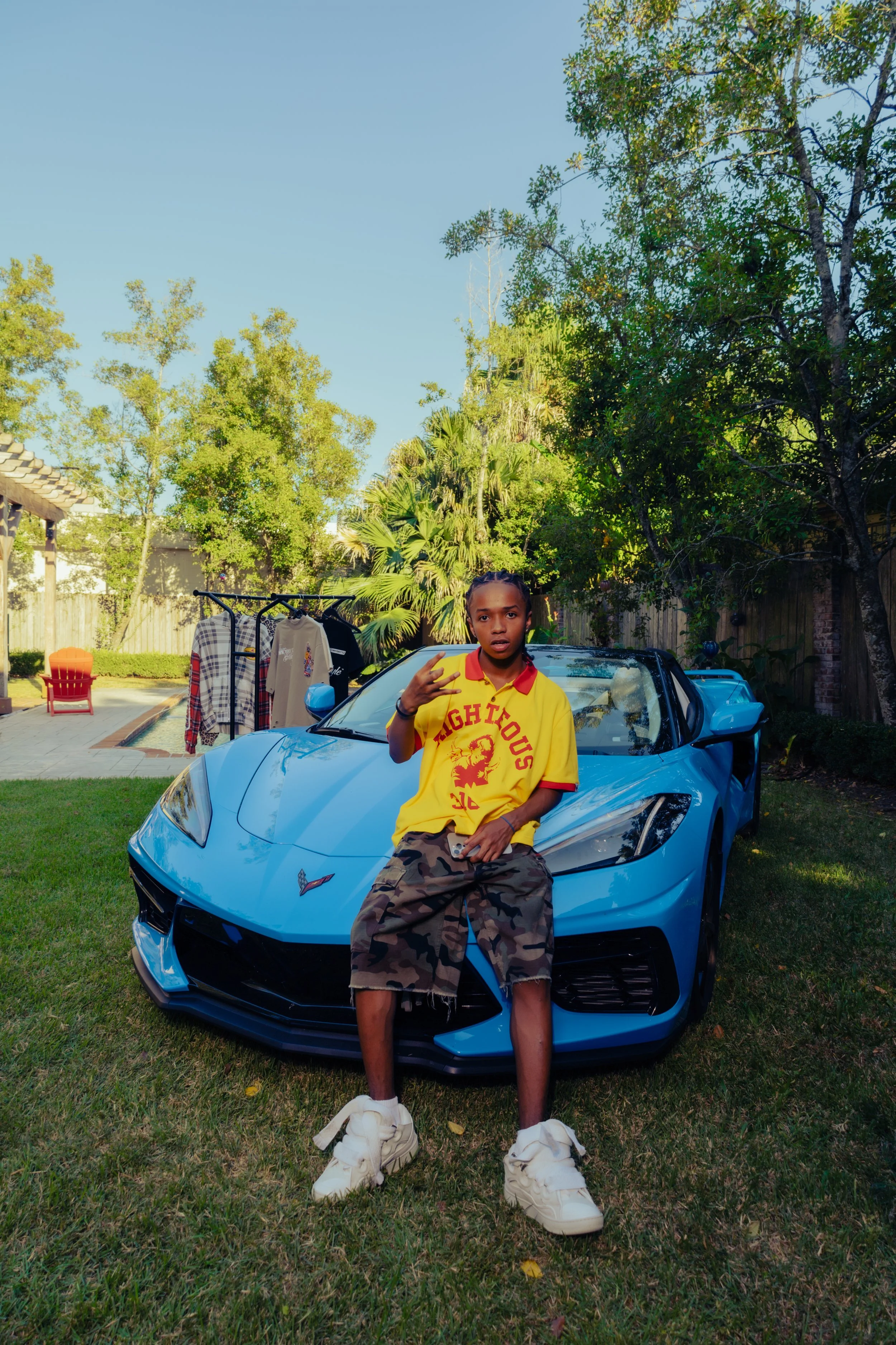 A young man wearing a yellow Toronto Raptors shirt and camouflage shorts is sitting on the hood of a blue sports car in a backyard, making a peace sign with his fingers. The background shows trees, a wooden fence, and outdoor furniture.