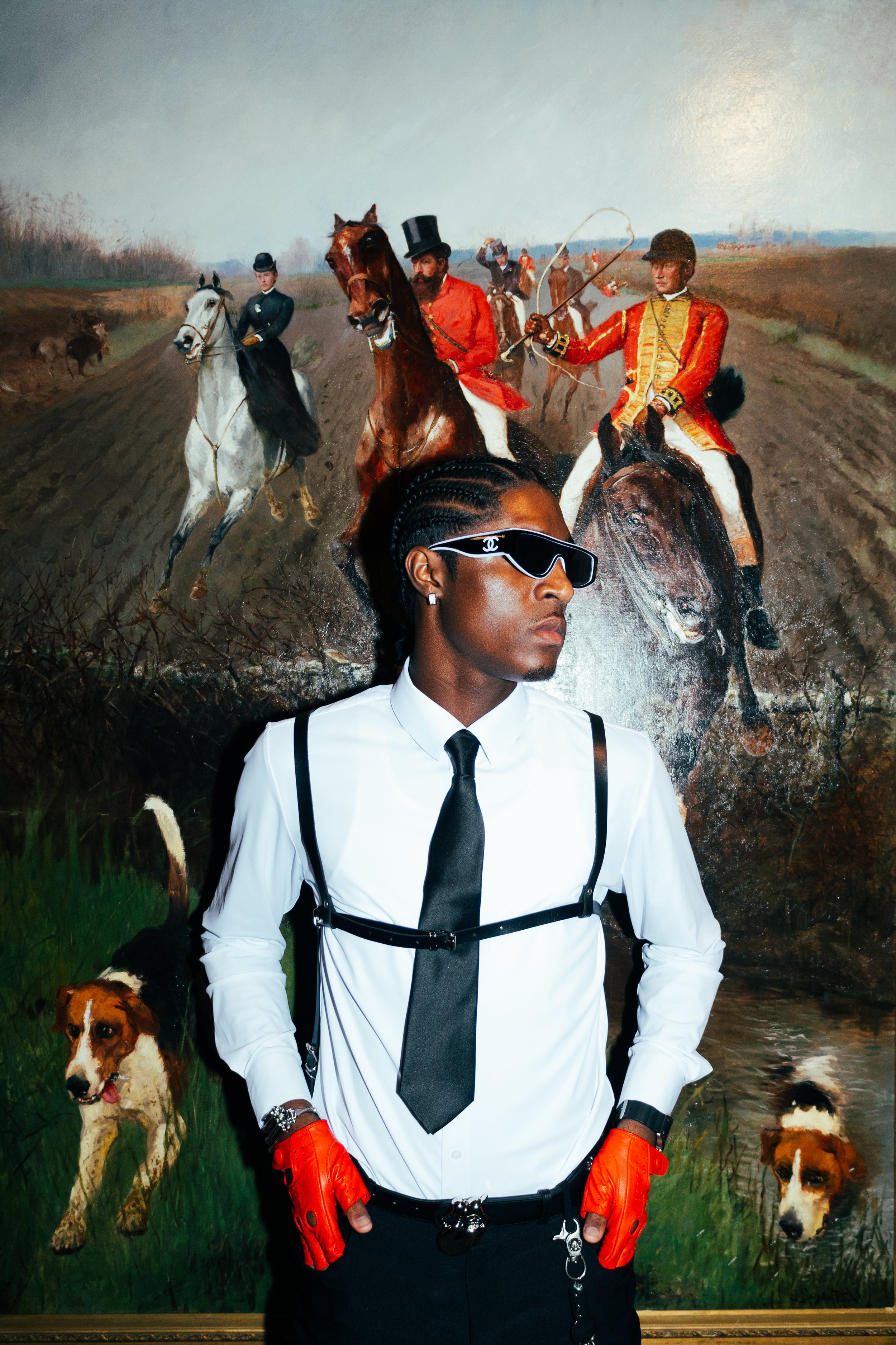 A person in a white shirt with black suspenders, wearing orange gloves, sunglasses, and a black tie, standing in front of a painting of a historical scene with men on horseback, dogs, and a landscape.