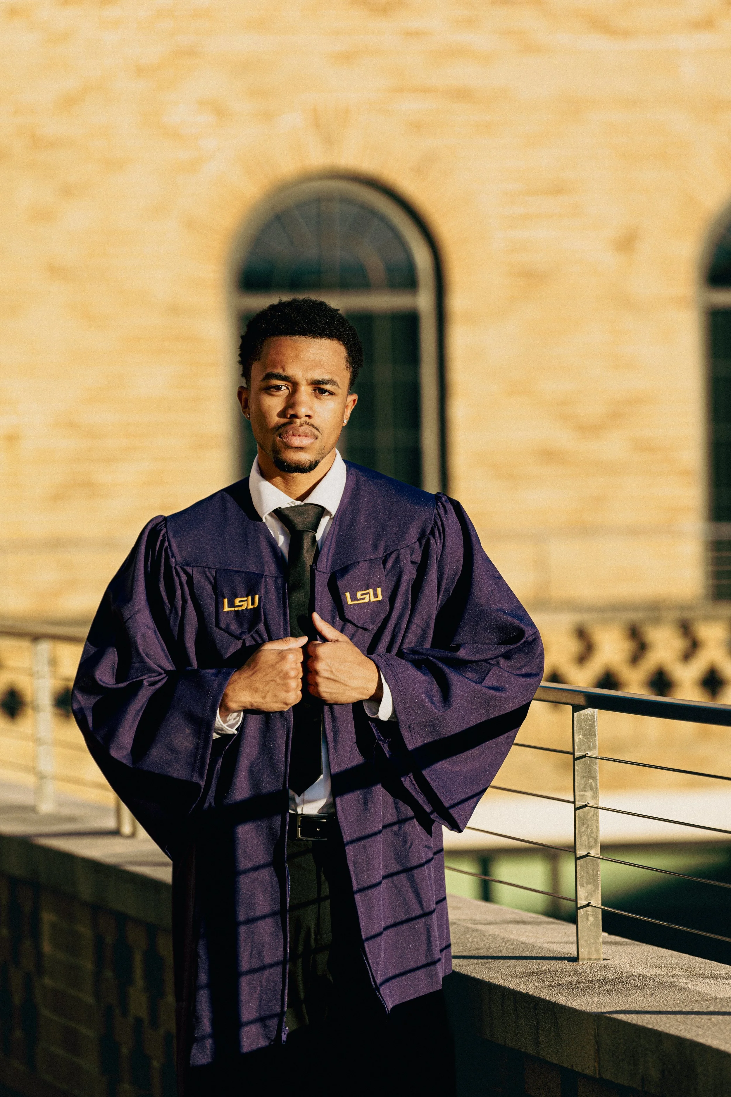 A young man in a graduation gown and cap standing outdoors near a railing, with a brick building and arched windows in the background, during sunset.