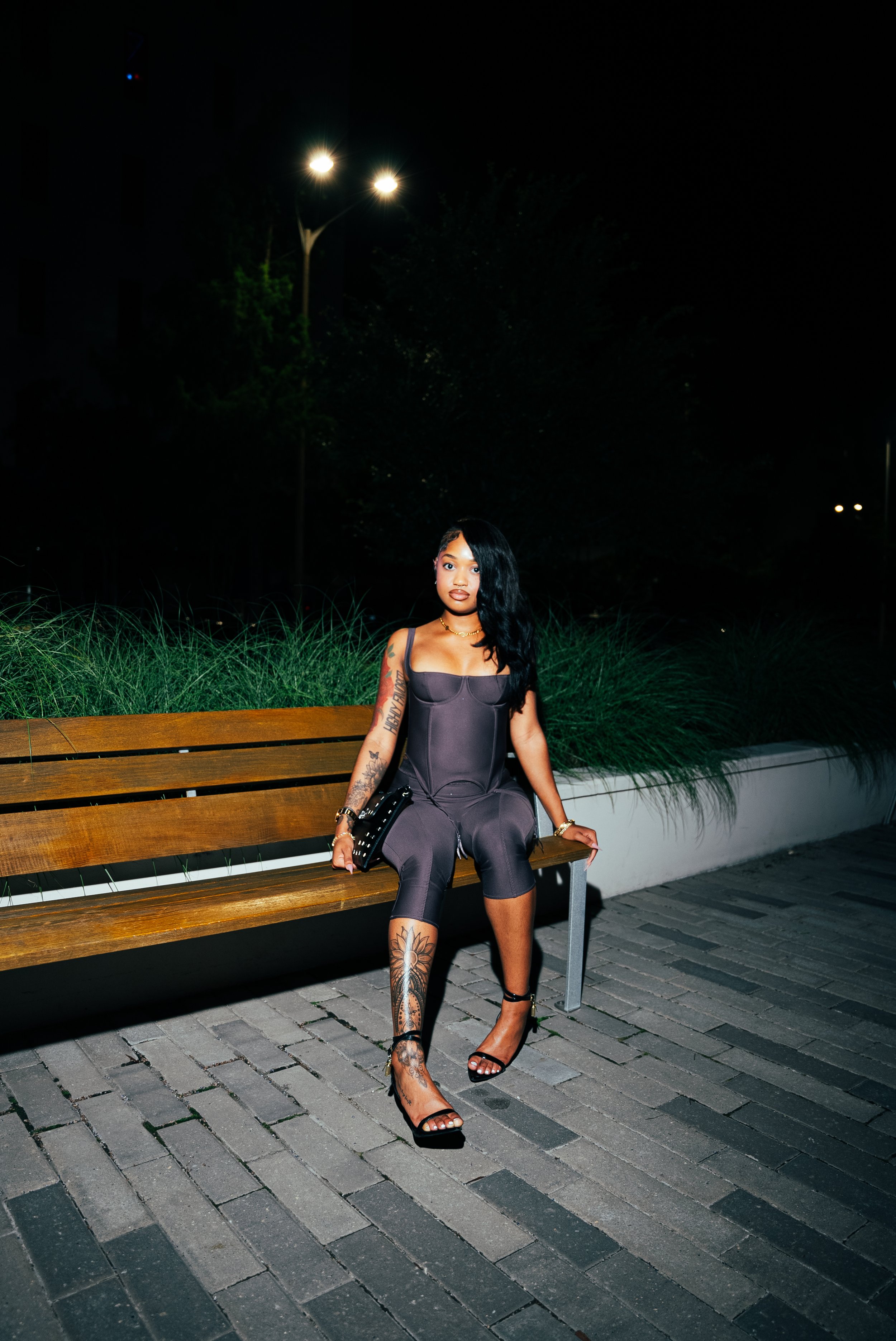 A young woman sitting on a wooden park bench at night. She has long black hair, tattoos on her left arm and right leg, and is wearing a dark fitted jumpsuit, high heels, and jewelry.