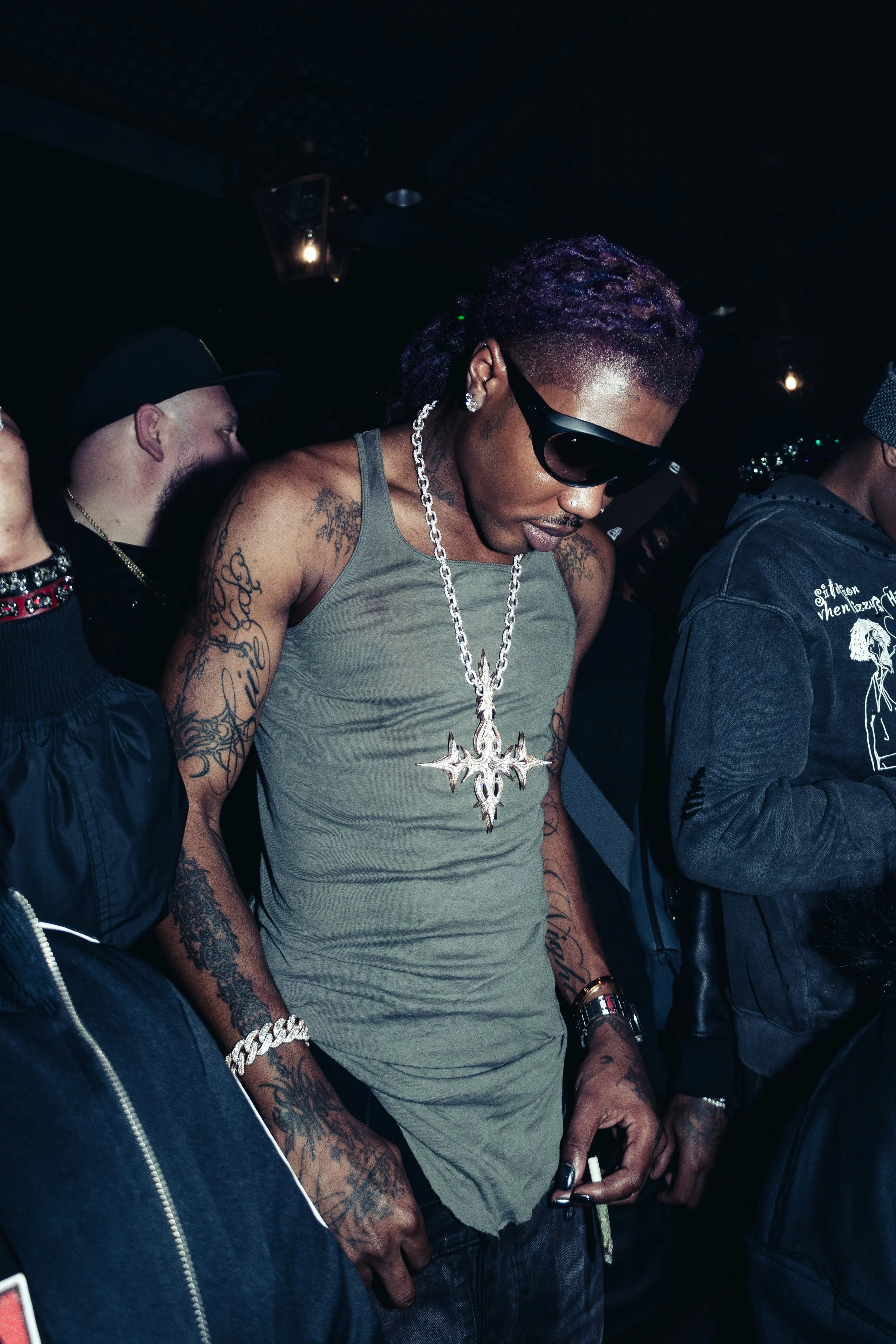 Young man with purple-tinted hair, large cross necklace, tattoos on arms, wearing sunglasses and a sleeveless shirt, in a dark, possibly club environment, with others nearby.