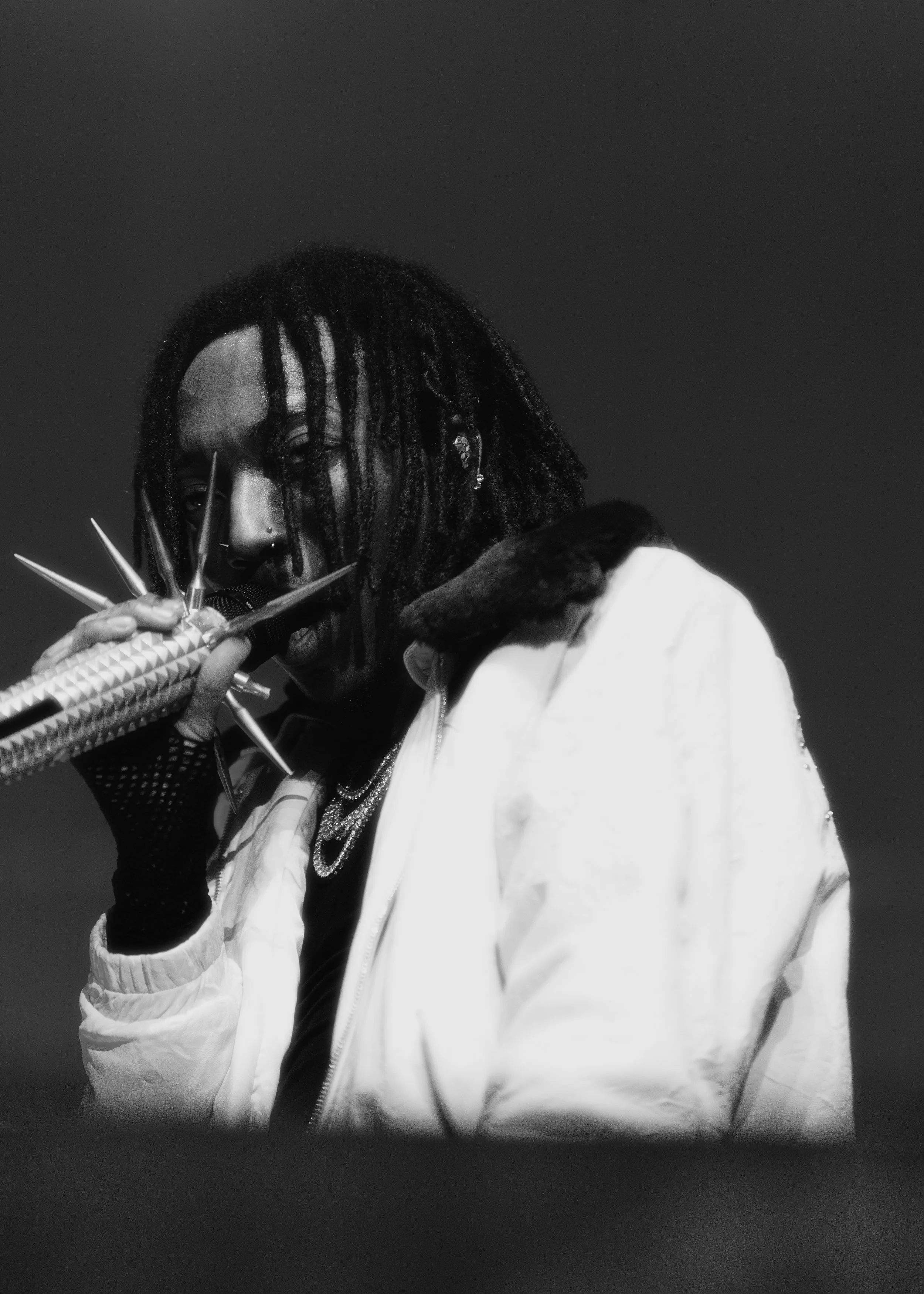 Black and white photo of a person with dreadlocks, holding multiple knives close to their face, wearing a white jacket with a fur collar.