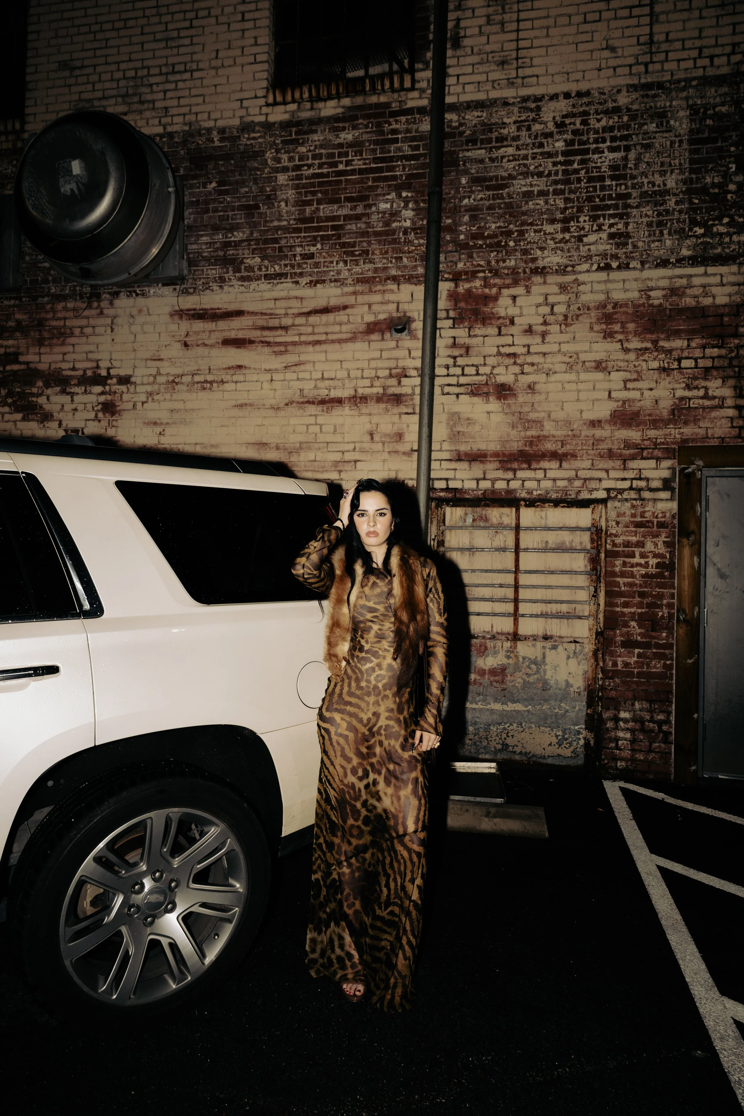 Woman in a long animal print dress and fur vest standing next to a white vehicle in a parking lot at night, with a brick wall in the background.
