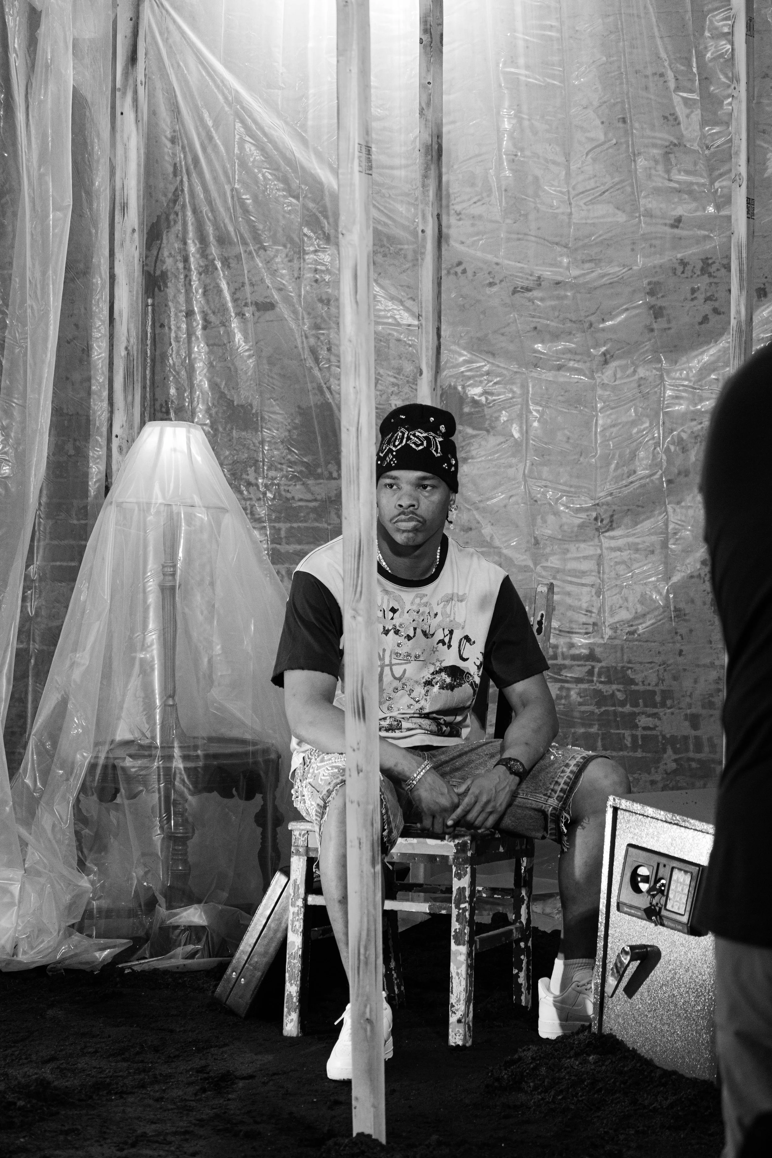 A young man sitting on a chair inside an unfinished room with plastic sheeting on the walls, wearing a beanie hat, a t-shirt, shorts, and sneakers, with a safe machine nearby.