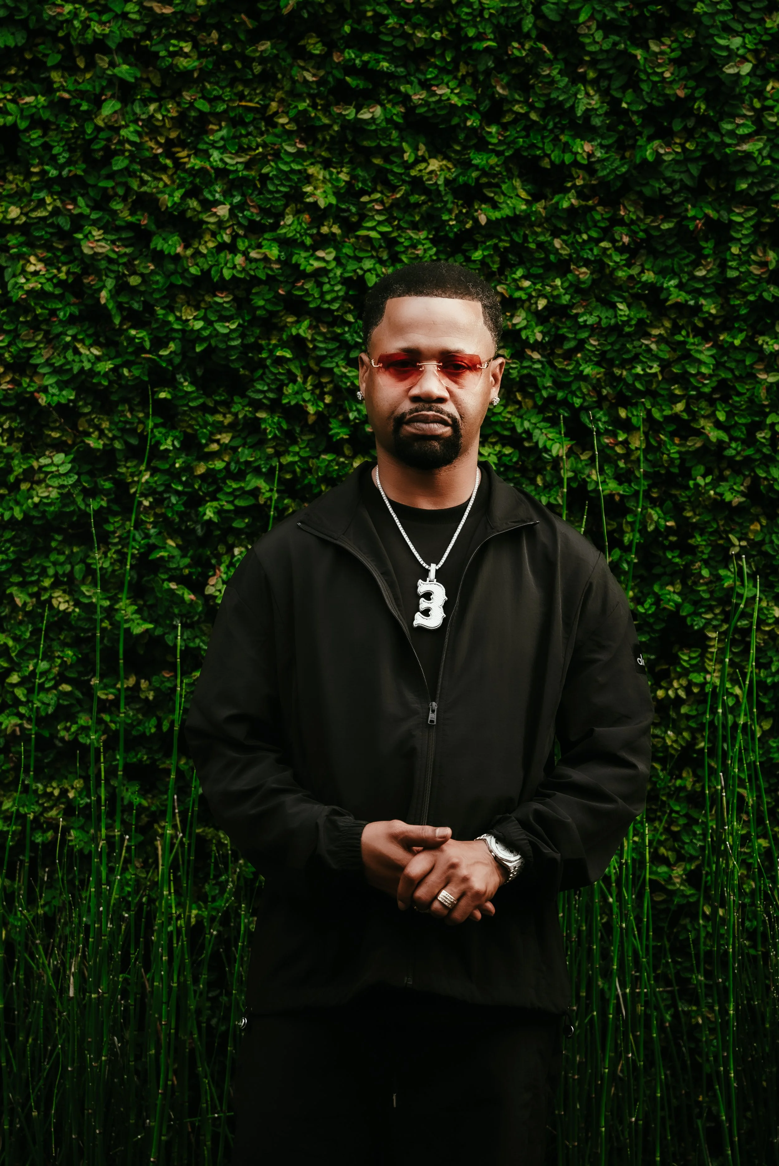 A man wearing red-tinted sunglasses, a black jacket, and a silver chain with a large pendant standing in front of a green leafy background.