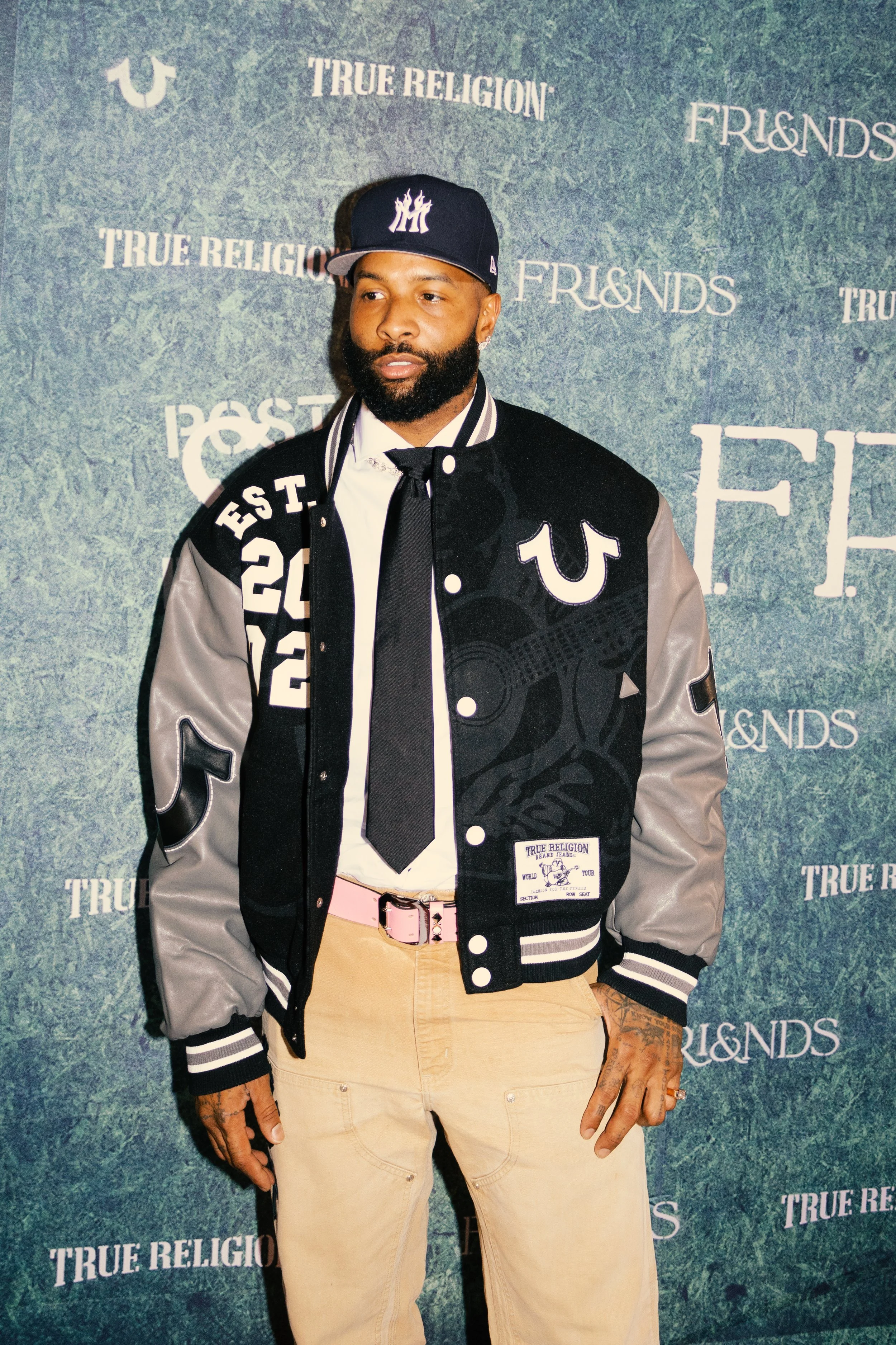 A man with a beard and tattoos on his hands wearing a black and grey varsity jacket, a white shirt, beige pants, and a black tie, standing in front of a blue-green textured backdrop with various words including 'TRUE RELIGION' and 'FRIENDS' printed o