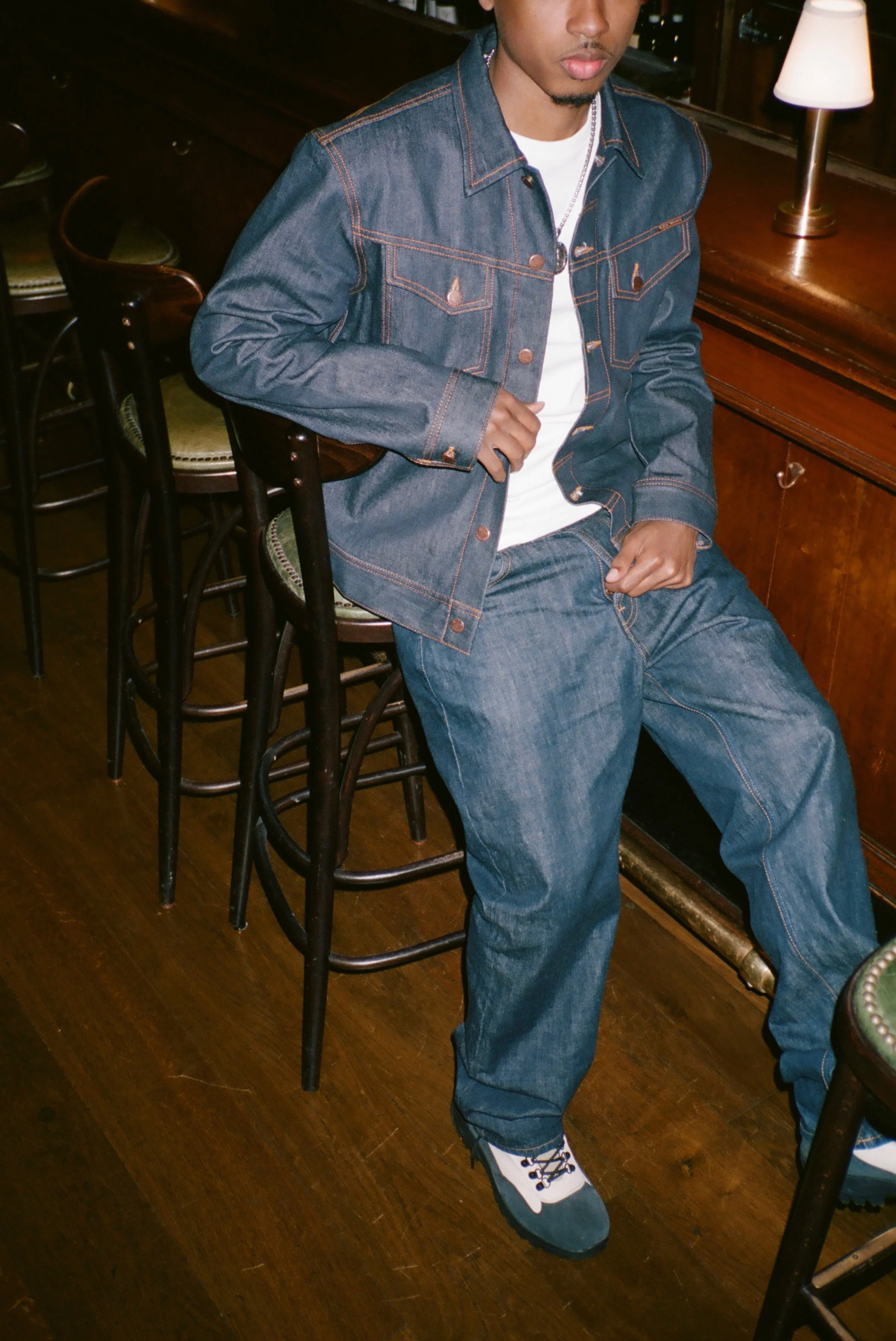 Young man dressed in a blue denim jacket and jeans, sitting on a barstool in a dimly lit room with wooden furniture and a lamp on the table.
