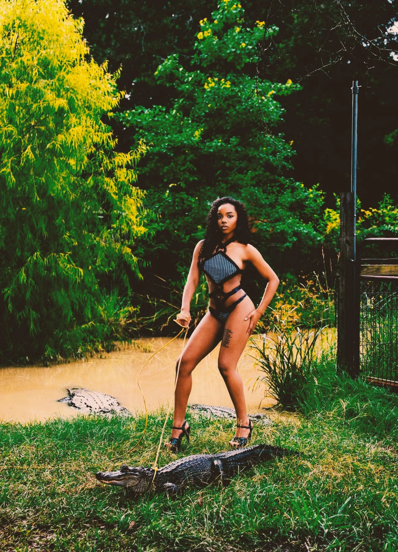 A woman with curly hair and tattoos on her thigh standing outdoors in a lush, green park near a crocodile. She is wearing a revealing black and white outfit and high heels, holding a leash attached to the crocodile's mouth. Surrounding trees are vibr