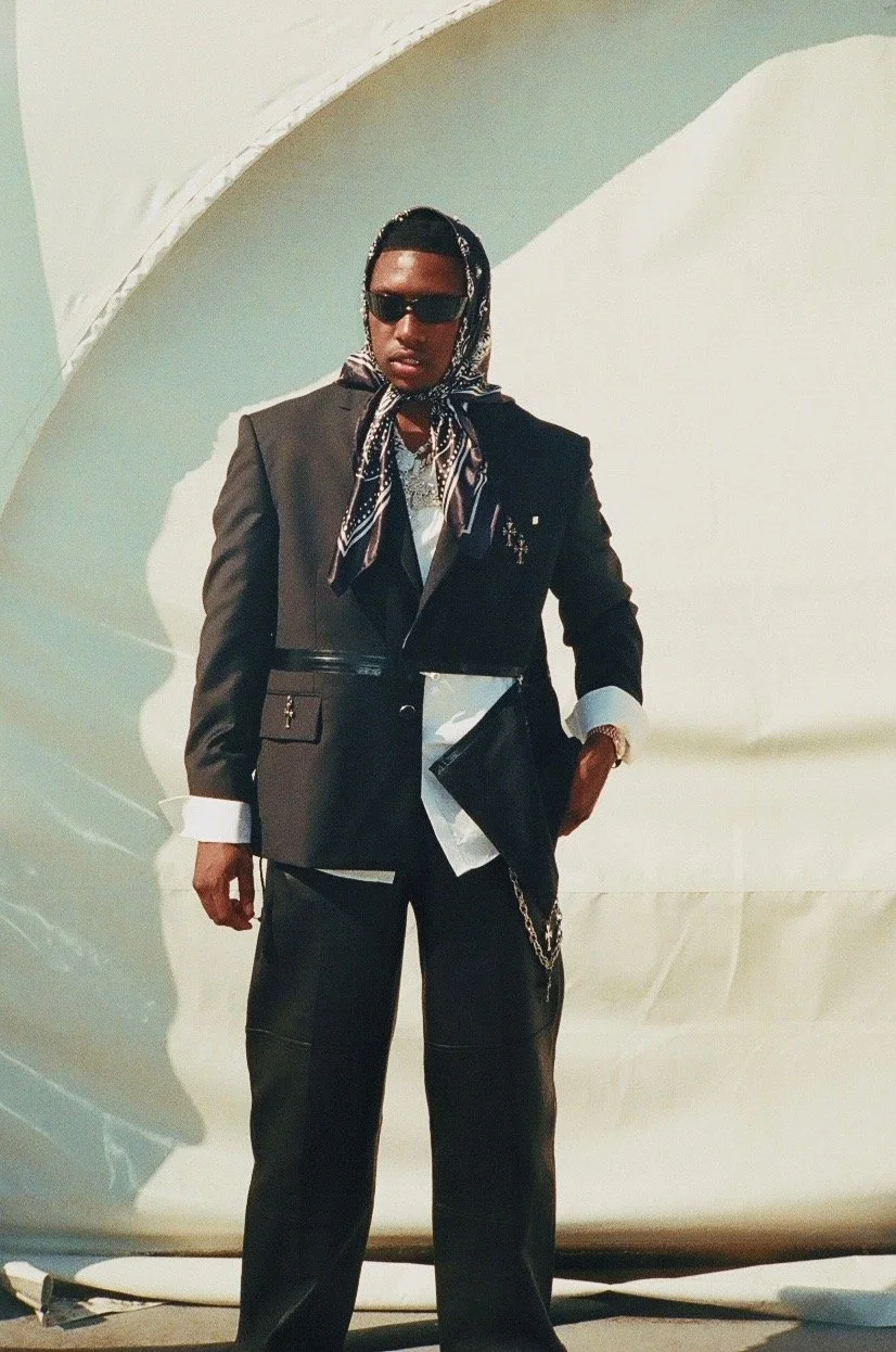 A person standing outdoors against a white curved wall, wearing a black blazer, black pants, a patterned scarf on their head, sunglasses, and jewelry.