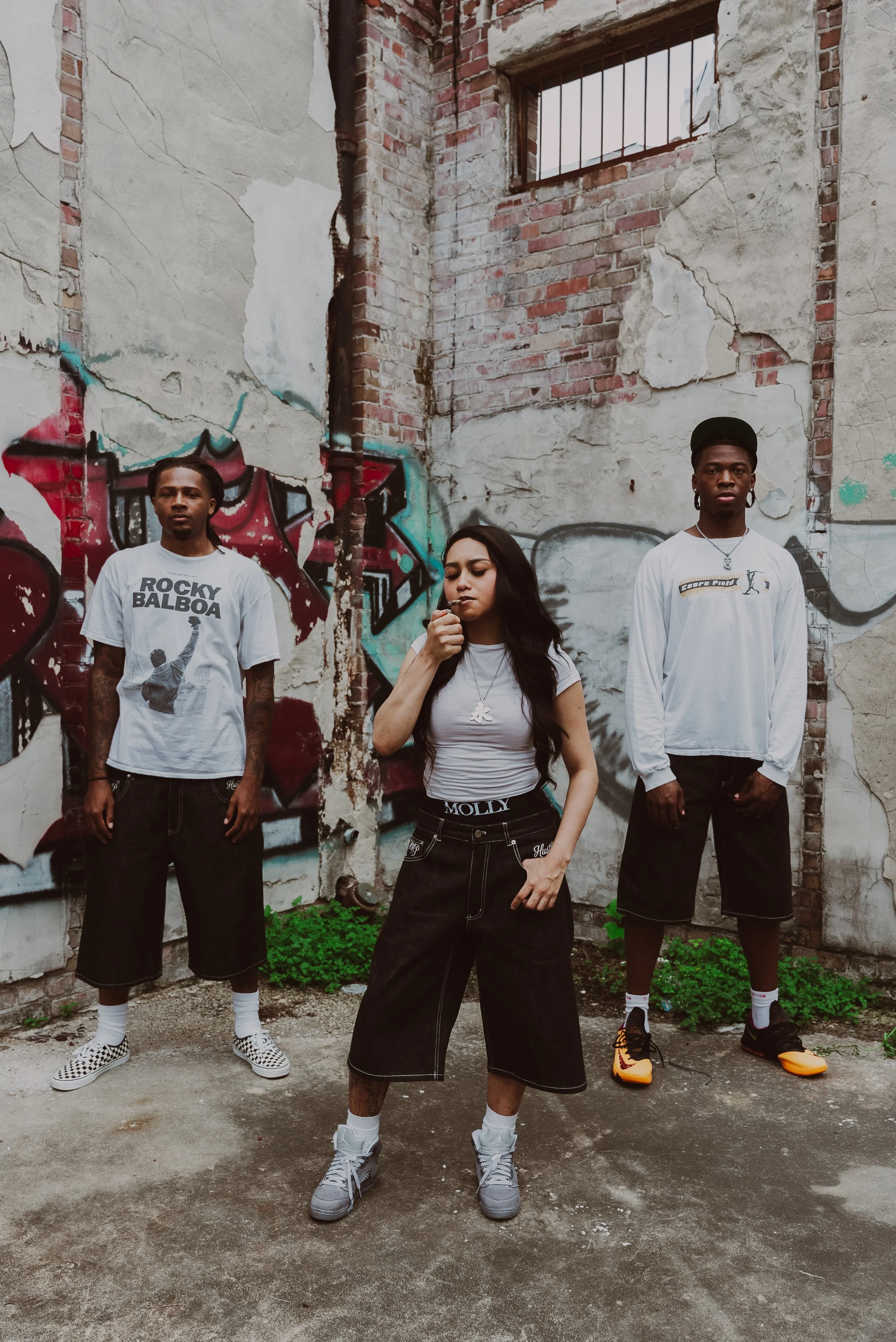 Three young adults standing in front of a graffiti-covered, weathered brick wall with a barred window. The person on the left wears a white Rocky Balboa t-shirt, black shorts, and checkered Vans sneakers. The person in the middle wears a white t-shir