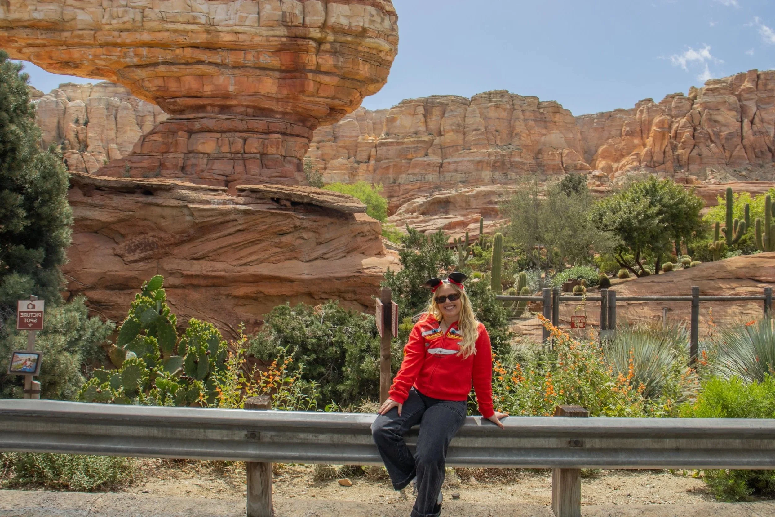 A woman with blonde hair, wearing sunglasses, a red jacket, and black pants, sitting on a guardrail in front of a desert landscape with large rock formations, cacti, and green shrubs under a partly cloudy sky.