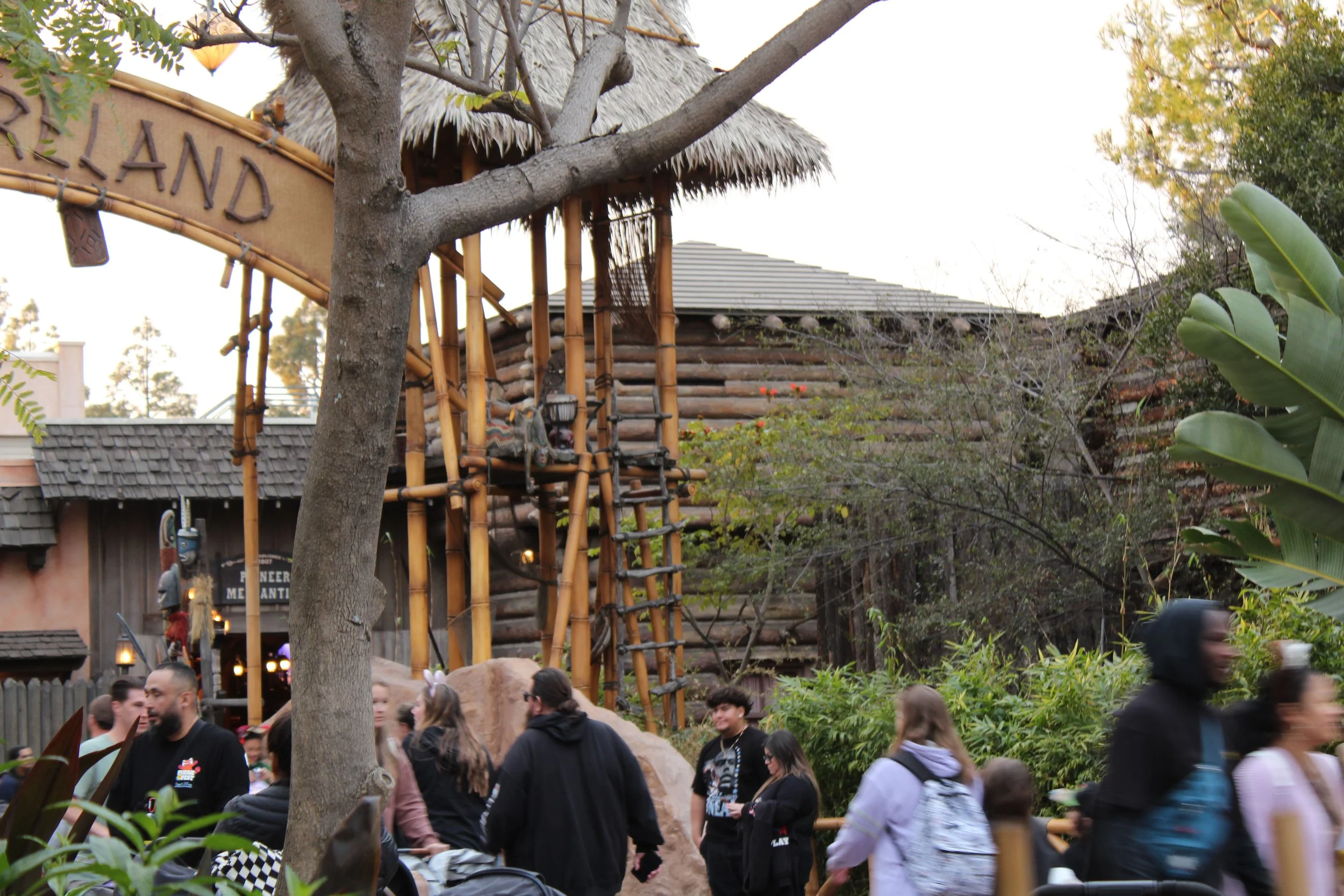 Theme park or amusement park scene with a wooden structure resembling a jungle gym or play area, surrounded by trees and lush greenery, with people walking and standing nearby.