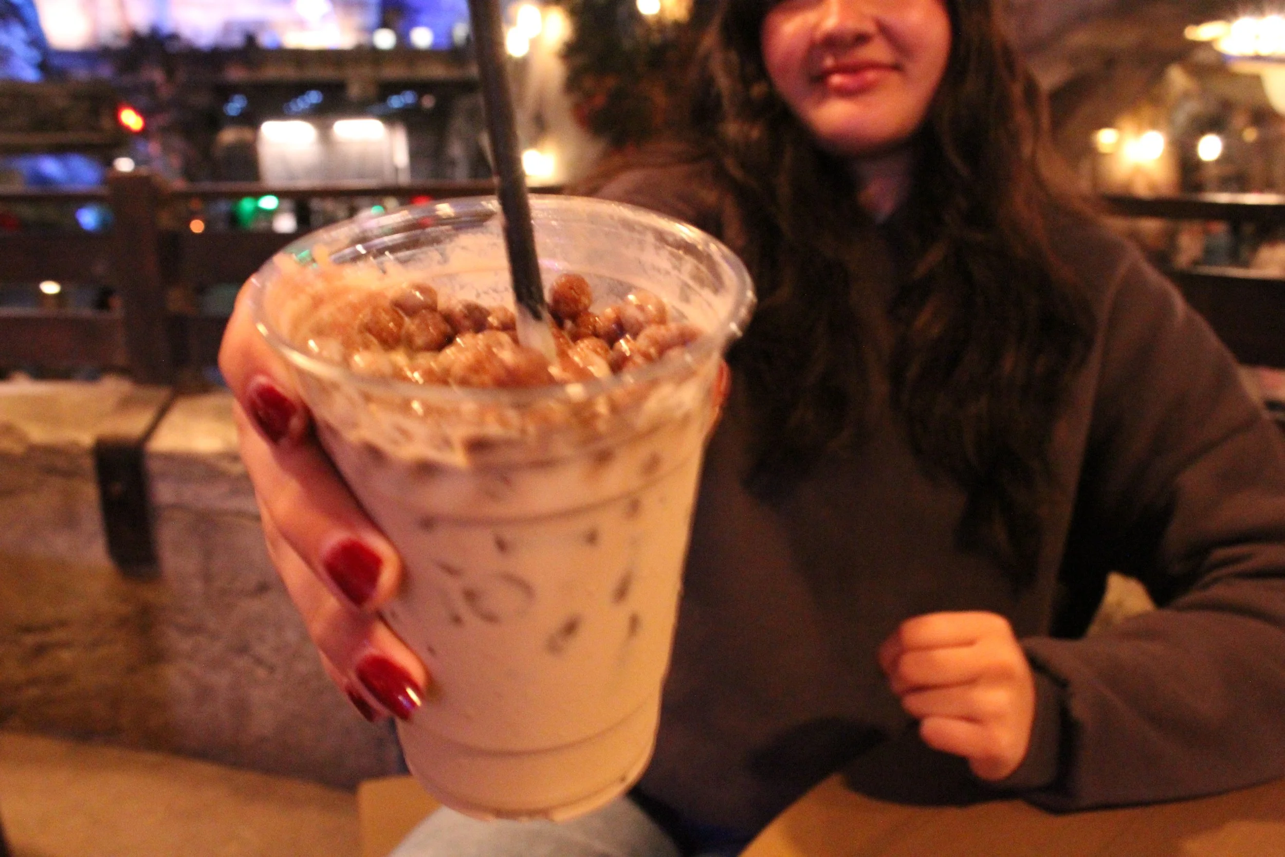 A woman with wavy brown hair and red nail polish holding a glass of chocolate milkshake topped with crumbled cookies or cereal in a restaurant or cafe setting.