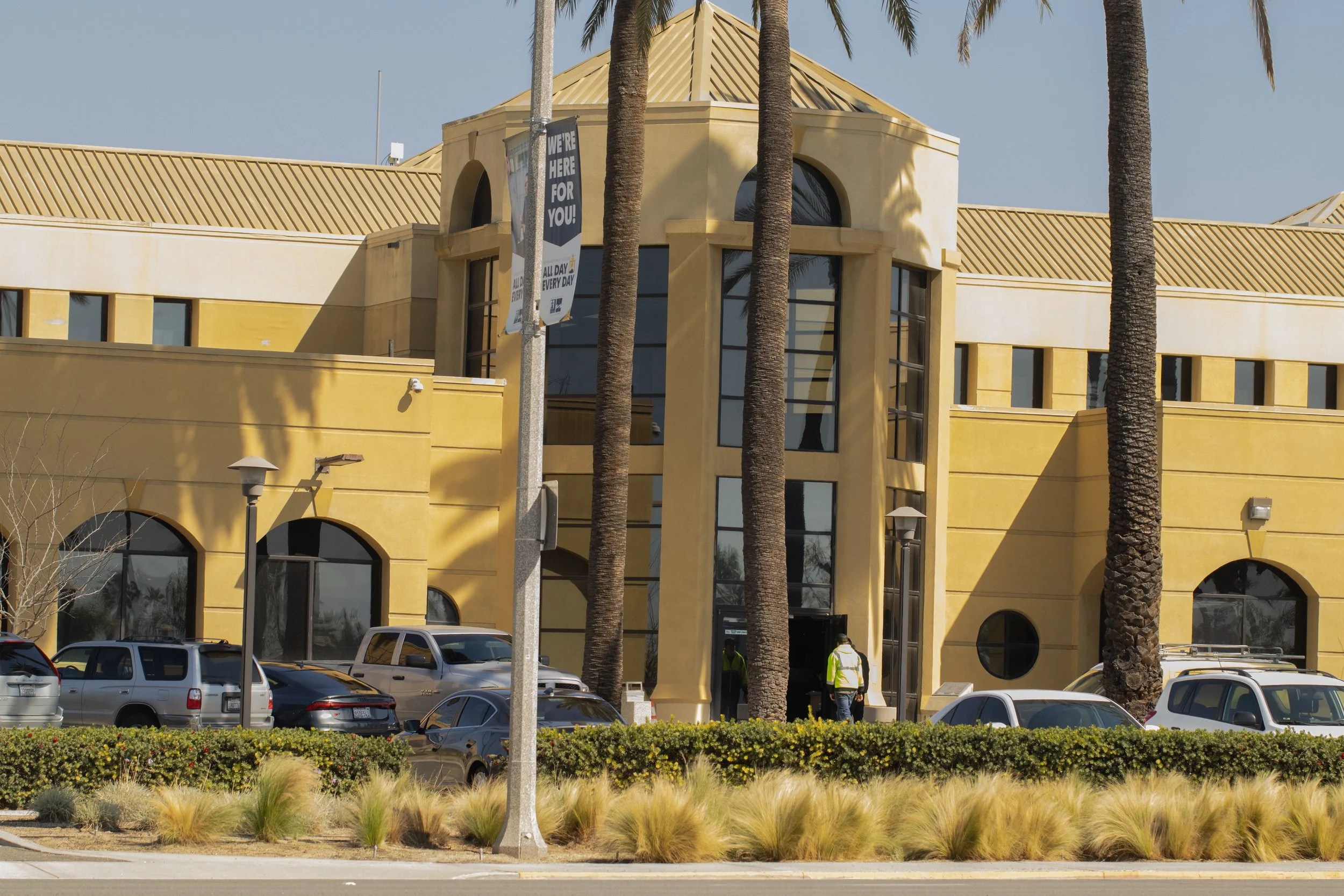 A large yellow building with arched windows and palm trees in front. Cars are parked outside, and there are people near the entrance.