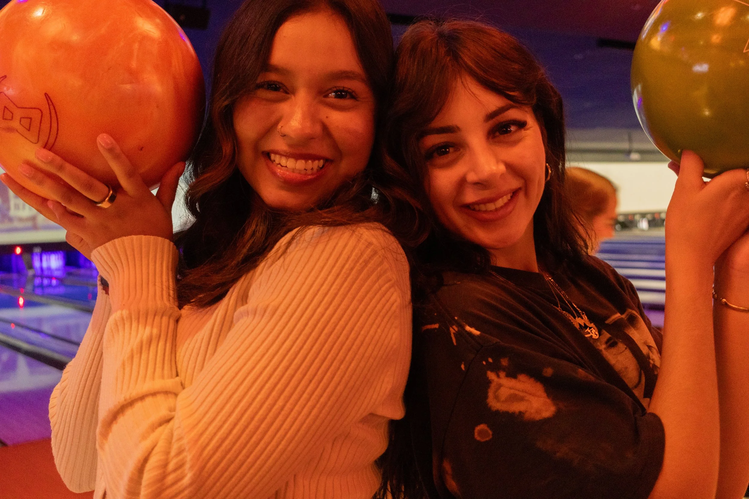 Two women smiling, holding colorful bowling balls in a bowling alley with dim lighting.