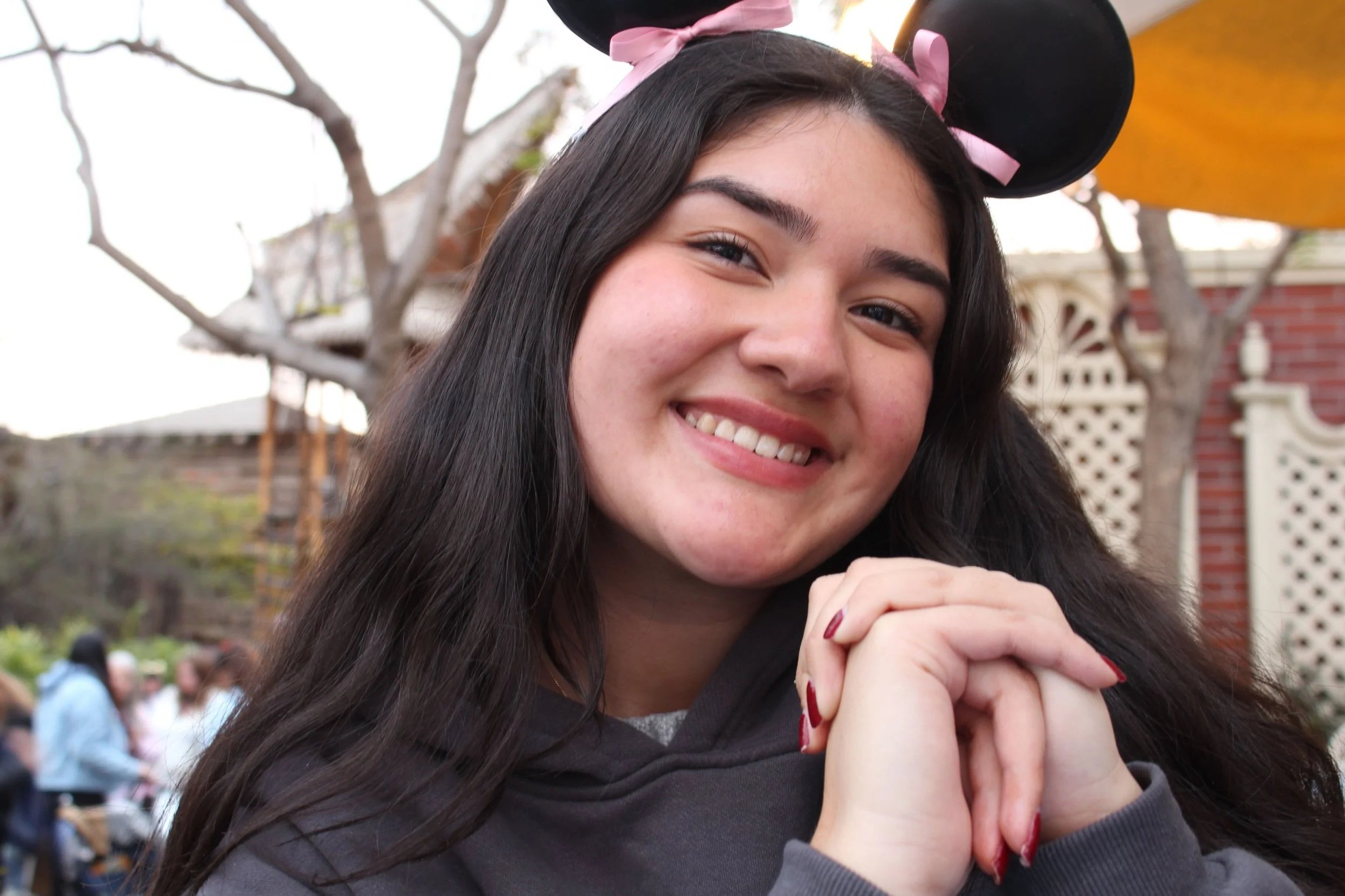 Young woman smiling with hands clasped, wearing Minnie Mouse ears with pink ribbons outdoors.