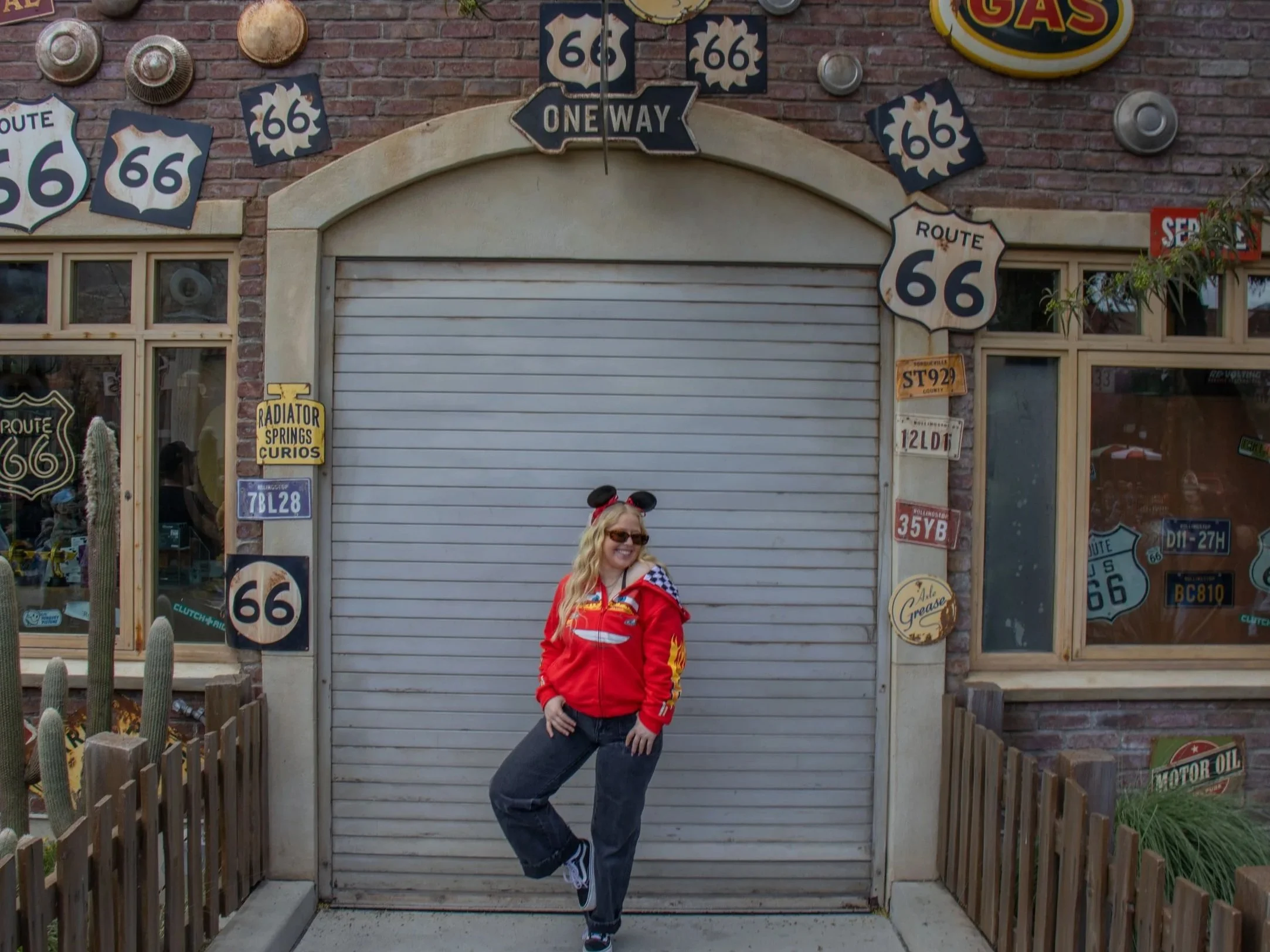 Woman with blonde hair wearing Mickey Mouse ears, sunglasses, red racing jacket, and jeans standing in front of a closed shop with Route 66 signs, license plates, and vintage signs on a brick wall.