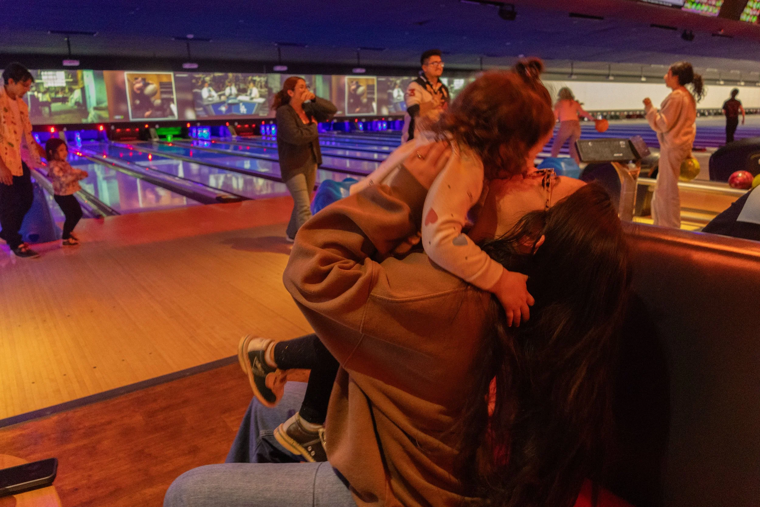 Two women are playfully fighting or hugging on the floor of a bowling alley, with other people in the background, some waiting for their turn to bowl and others watching. The bowling alley has colorful lighting and multiple lanes.