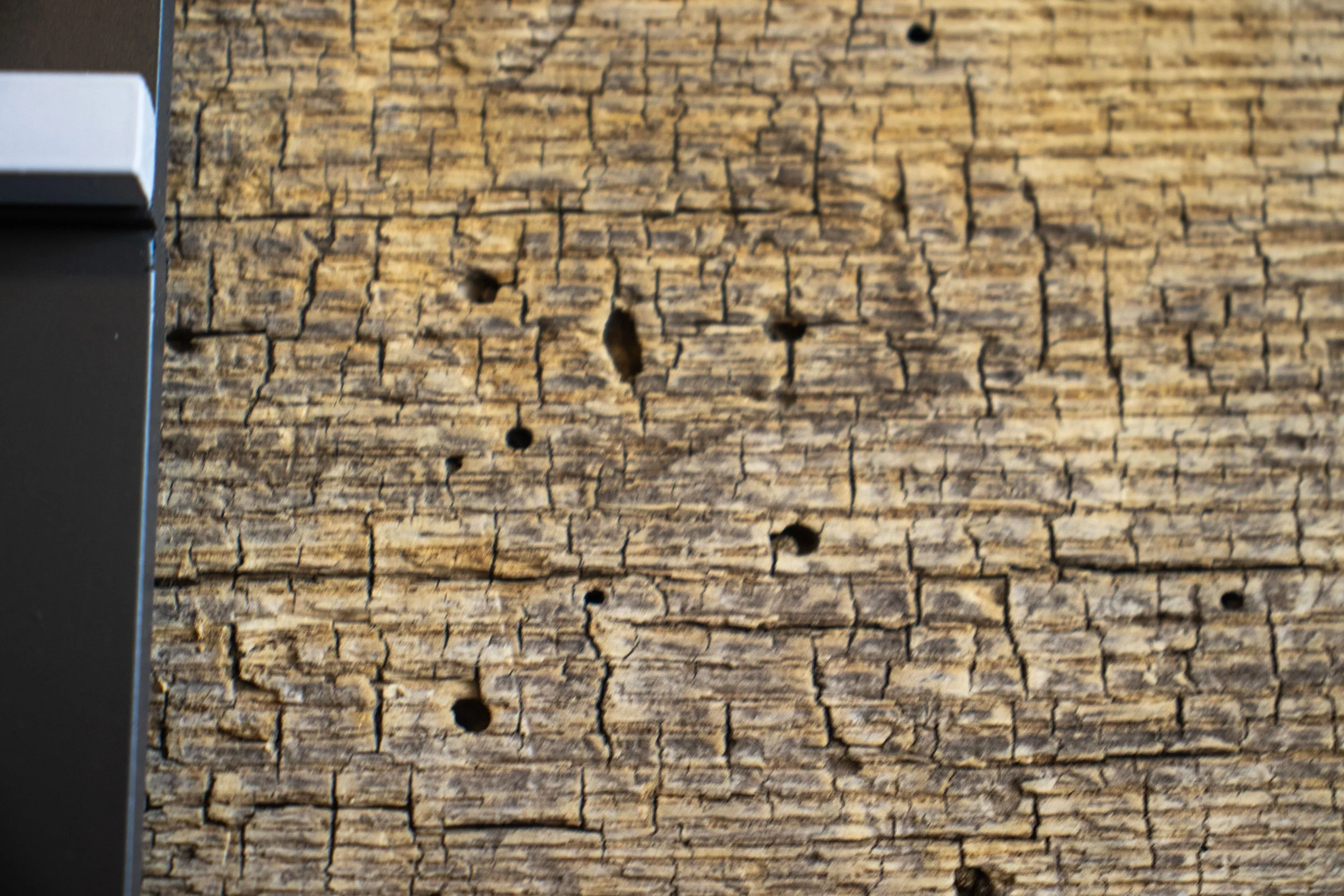 Close-up of a weathered wooden surface with visible cracks and holes, partially showing a dark gray or black machine or object on the left side.