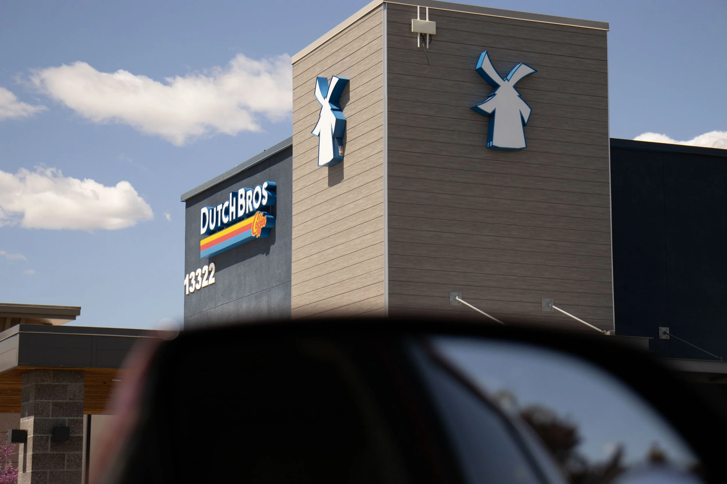 The exterior of a Dutch Bros coffee shop with the Dutch Bros logo and the number 13322, featuring a gray and beige building with a large windmill logo on the side. Part of a car's side mirror is visible in the foreground.