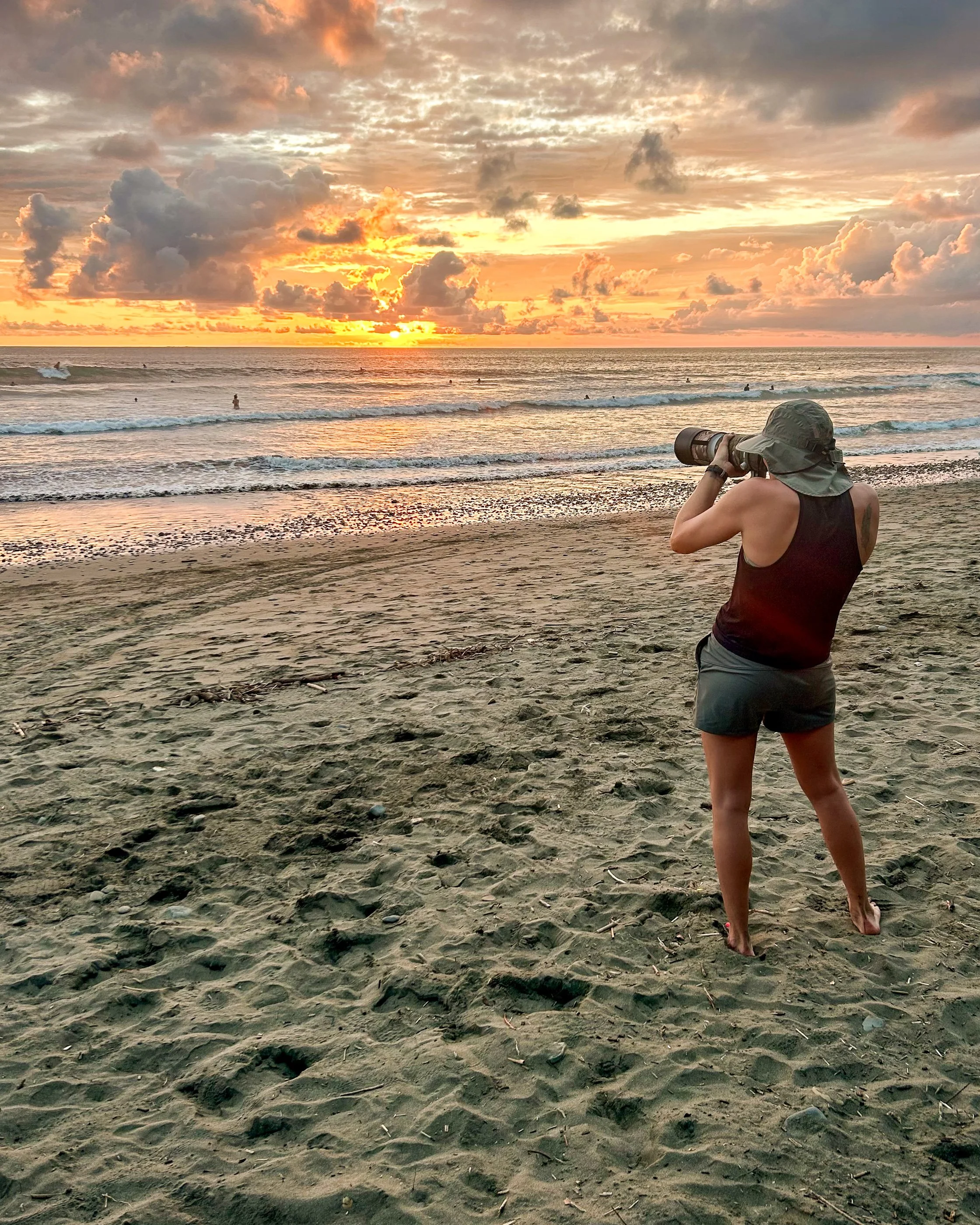A person standing on a sandy beach, wearing a hat, black sleeveless shirt, and shorts, taking a photo with a camera, with a sunset over the ocean in the background.
