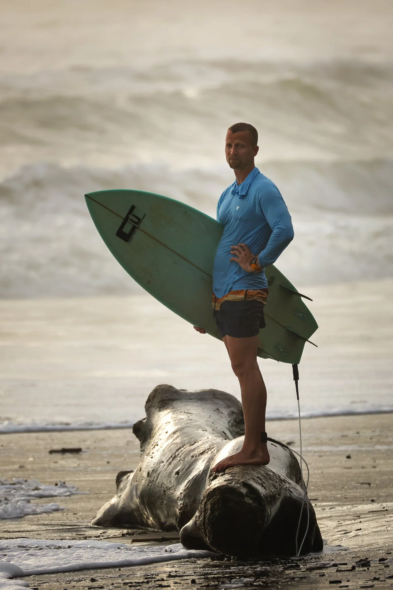 A man standing on a fallen log at the beach, holding a surfboard, with ocean waves in the background.