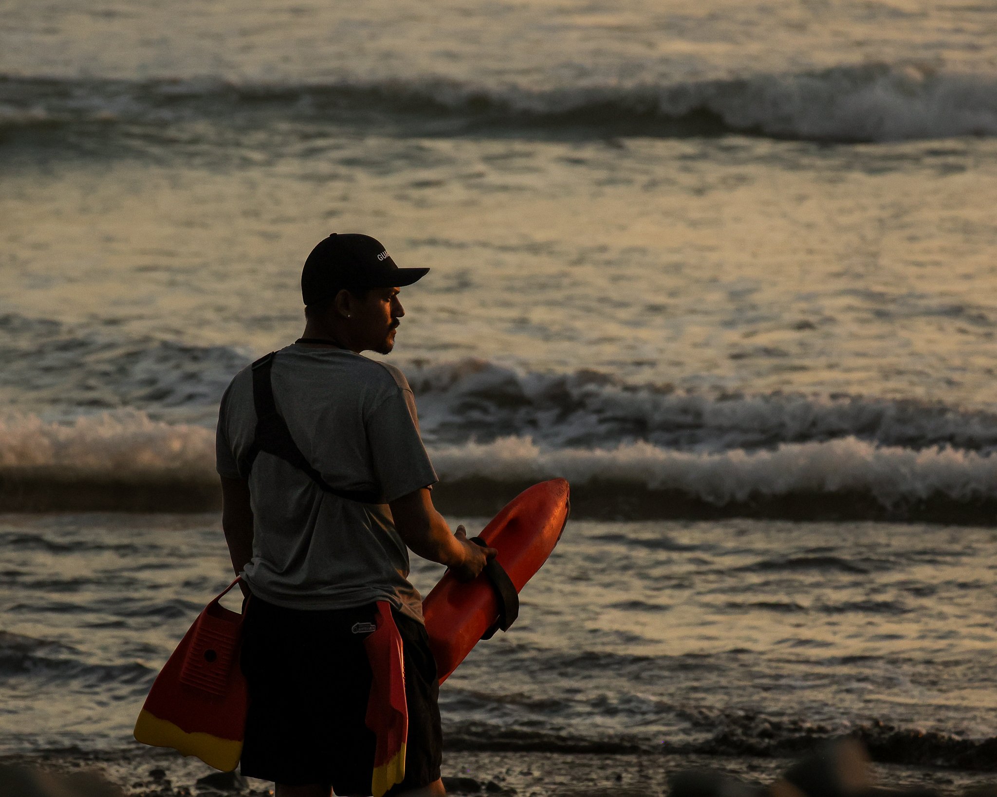 A lifeguard standing on the beach holding a rescue float, wearing a cap and casual clothes, with ocean waves in the background during sunset.