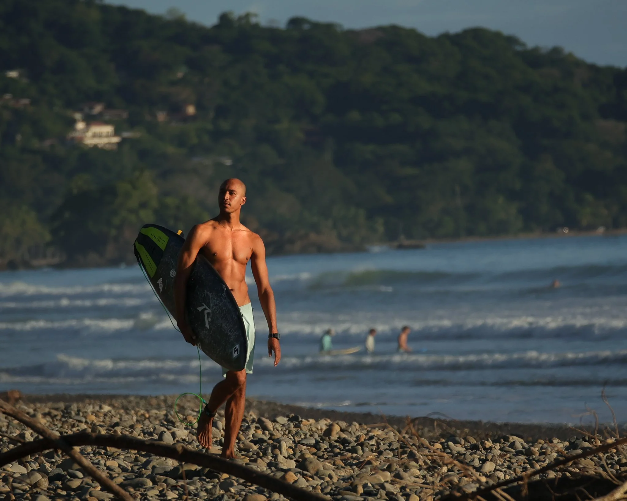A shirtless man holding a surfboard walking on a rocky beach with waves and other surfers in the background.