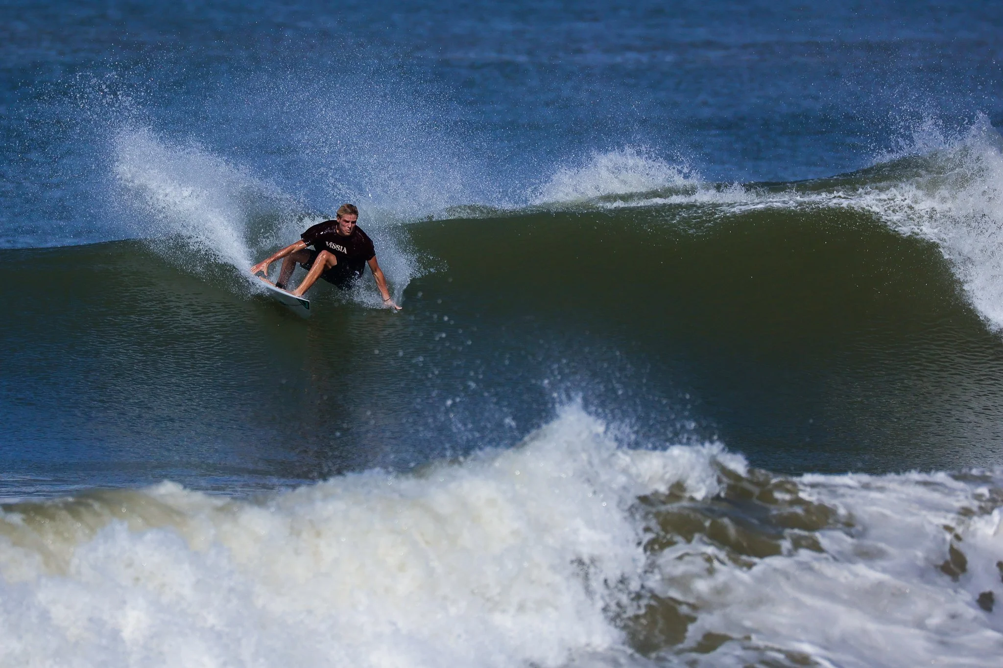 A male surfer rides a large wave on a surfboard, wearing a black shirt, with water splashing around him.
