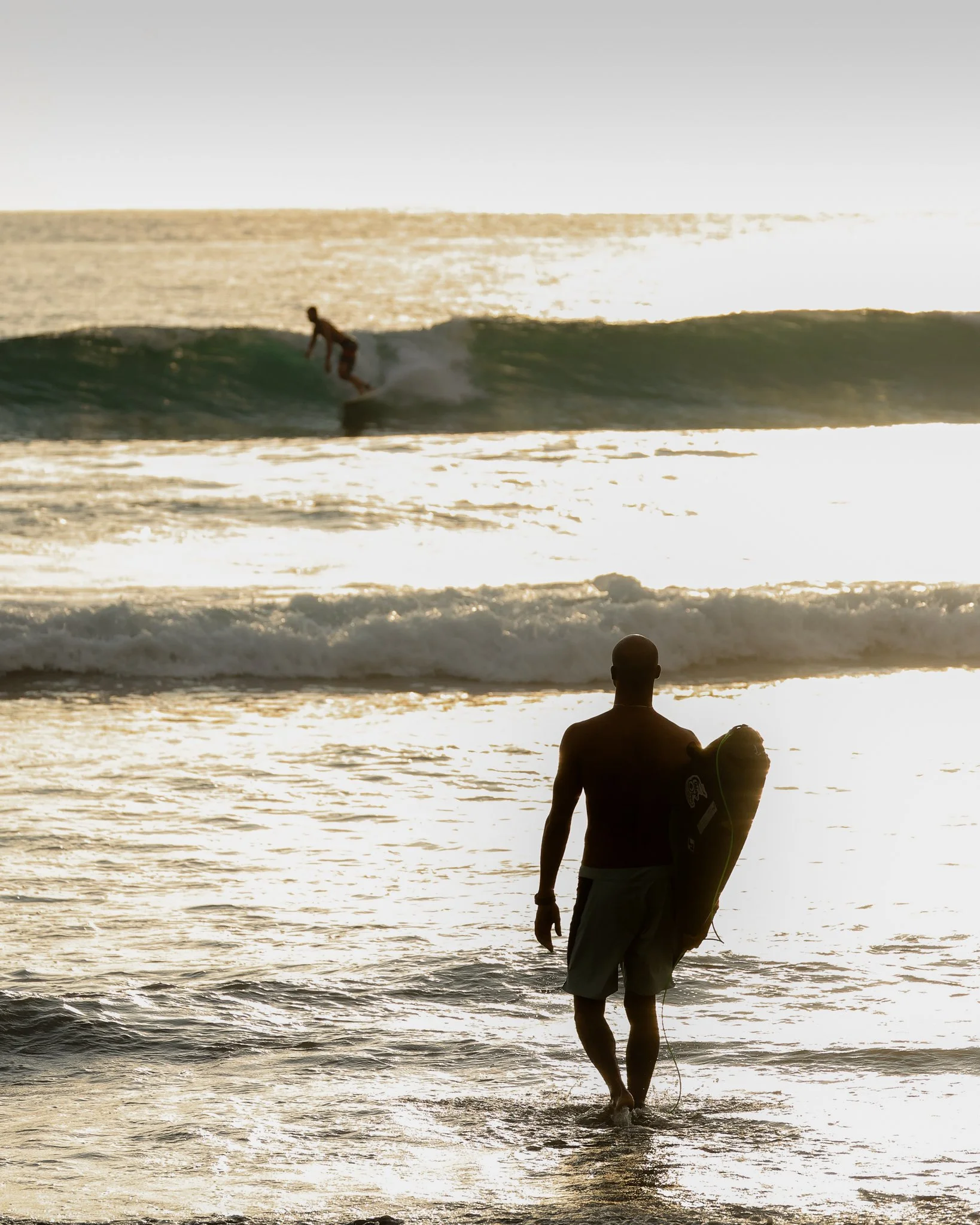 A person walking into the water carrying a surfboard while another person surfs a wave in the background at sunset.