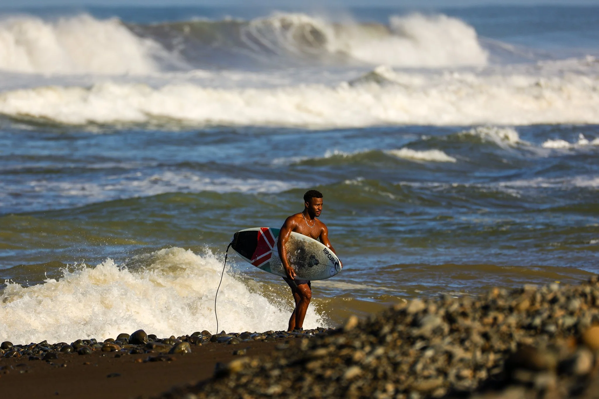 A man with a surfboard standing on a rocky beach with ocean waves in the background.