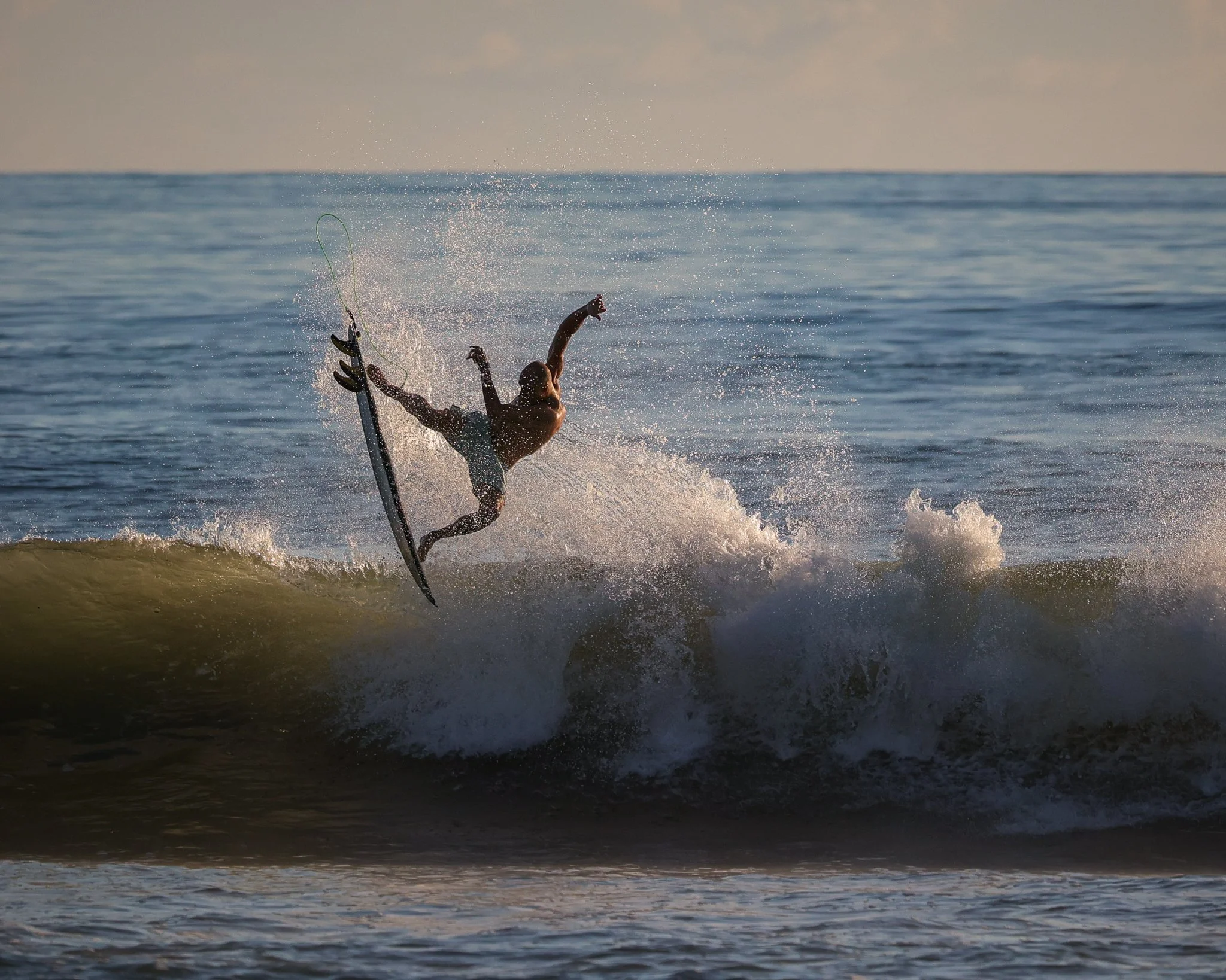 A person performing an aerial trick on a surfboard over a breaking wave at the beach during sunset.