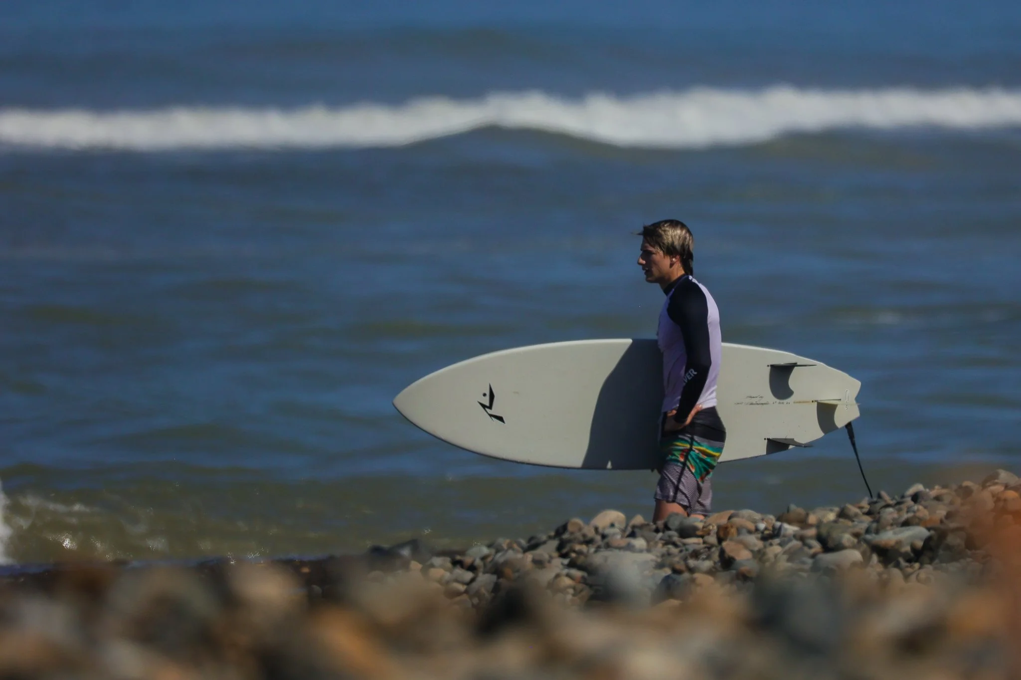 Man walking on rocky beach while holding a surfboard, with ocean waves in the background.