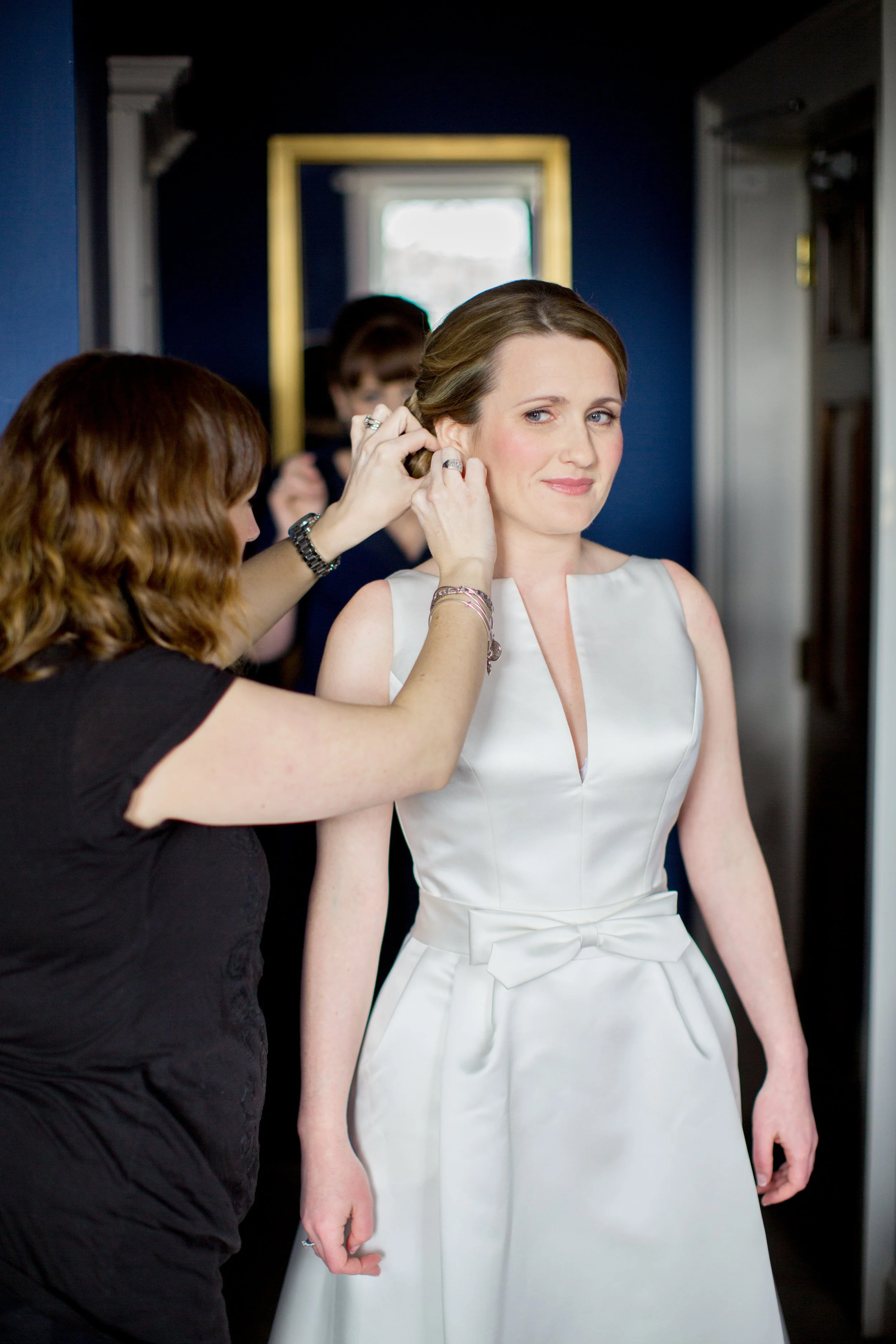 A woman in a white dress is being dressed in earrings by another woman, with her reflection visible in the mirror behind them.