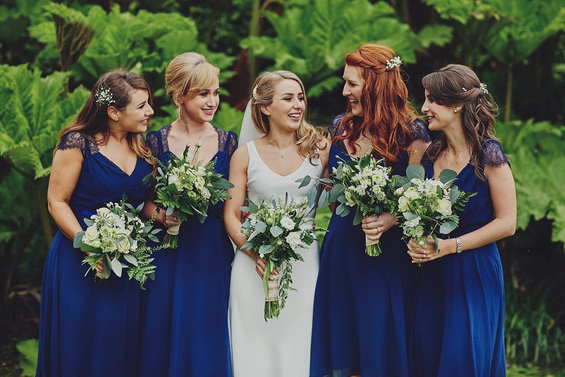 A bride and five bridesmaids standing outdoors on a lush green background, all holding bouquets of white flowers and greenery. The bridesmaids are wearing matching dark blue dresses with lace sleeves, and the bride is in a white wedding gown.