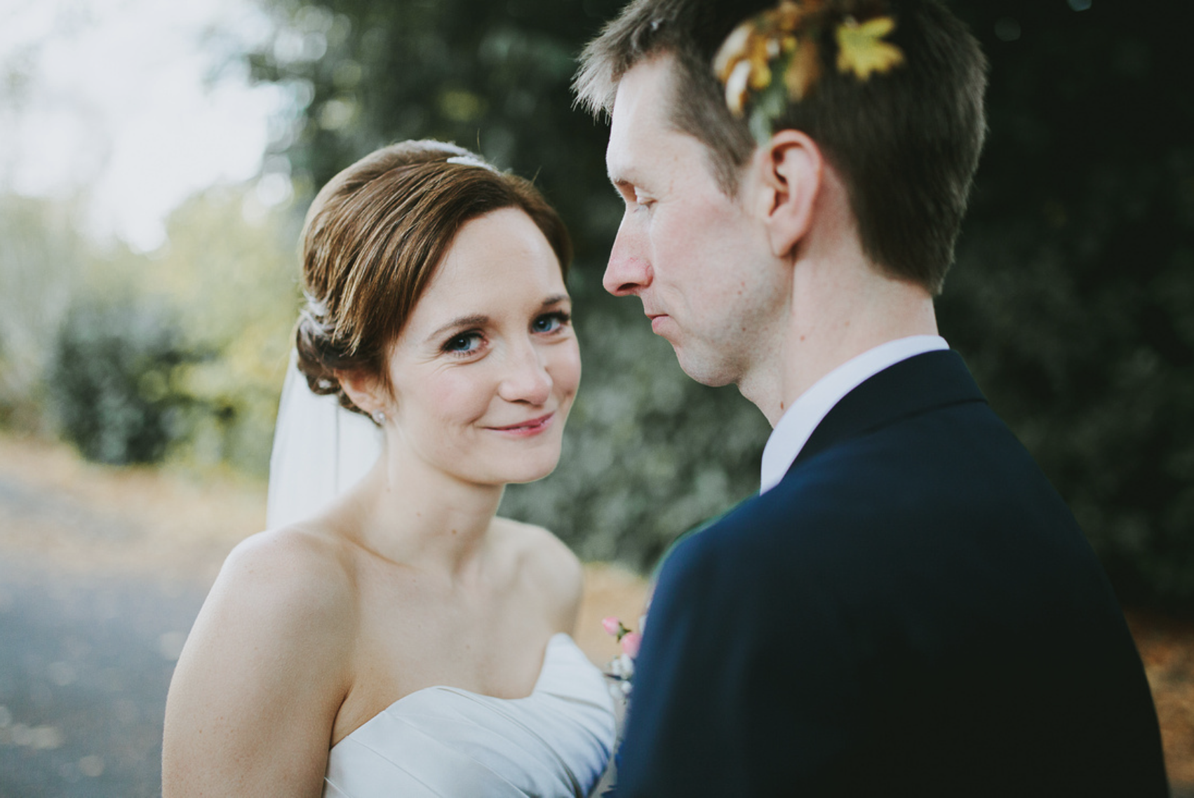 A bride and groom face each other closely outdoors, with the bride smiling softly and the groom looking serious. The bride has short, wavy auburn hair styled neatly, wearing a strapless white wedding dress and a veil, while the groom has short brown 